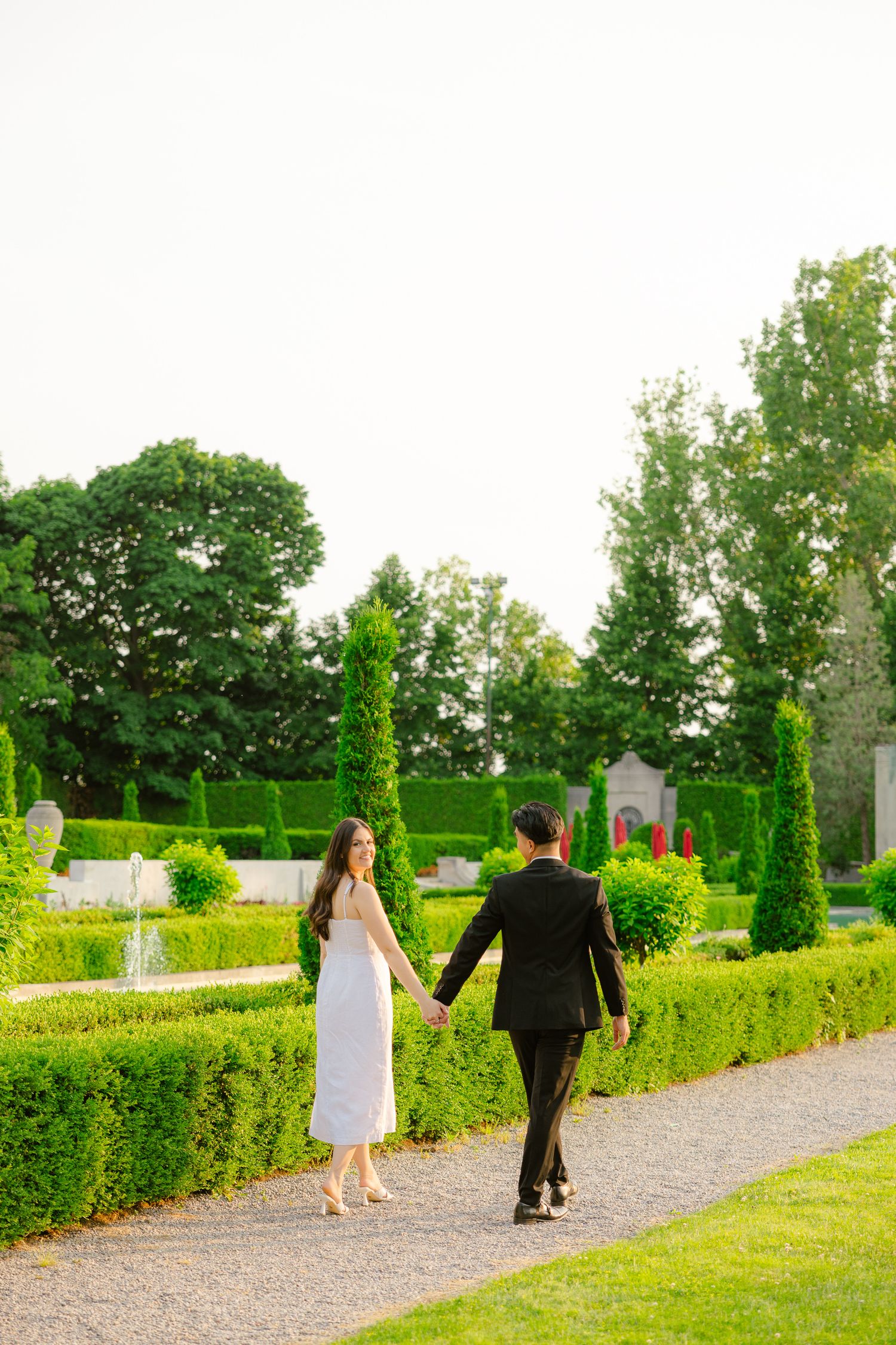 A couple holding hands while walking through a lush garden with perfectly trimmed topiaries.