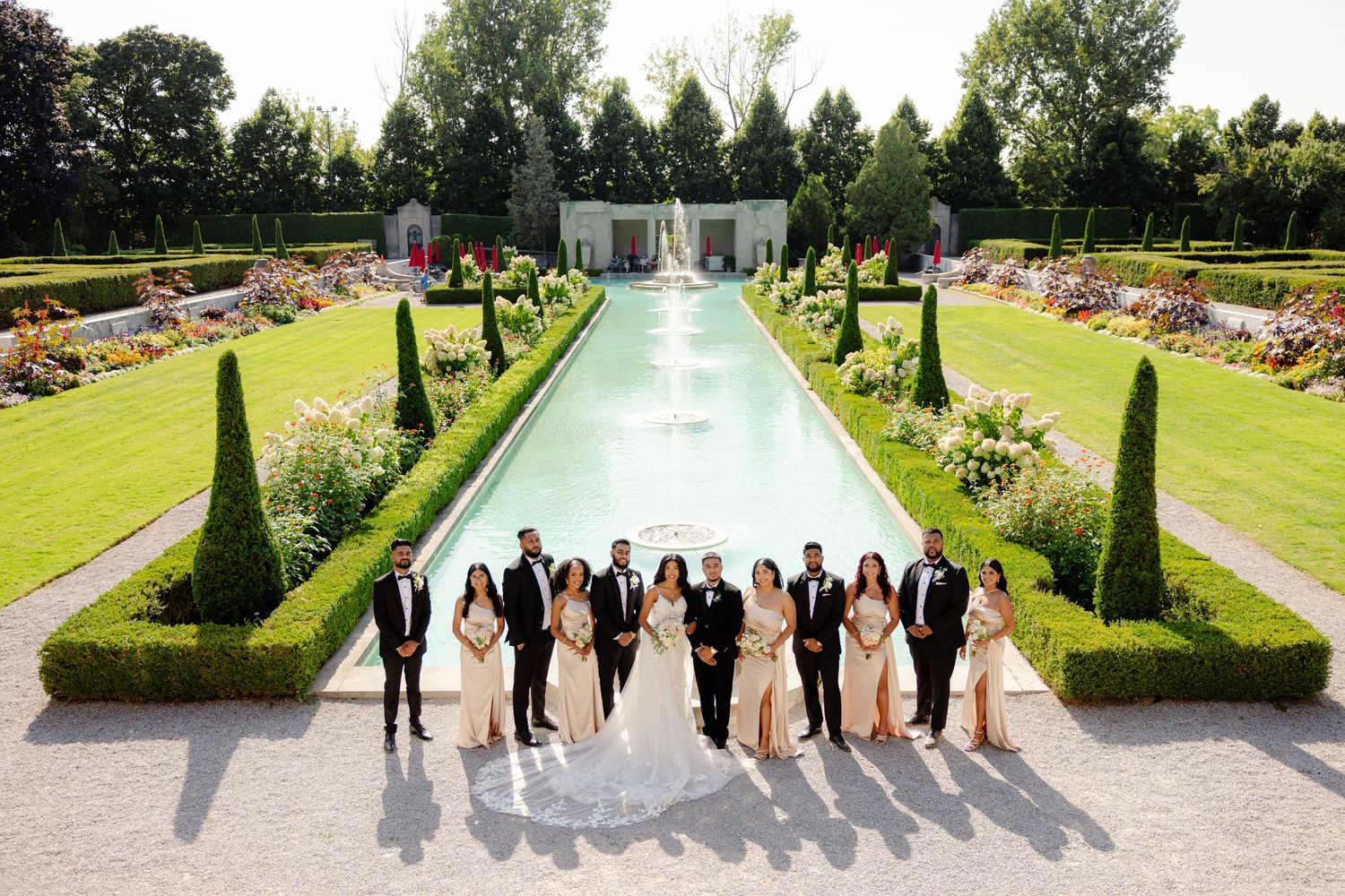 Bridal party poses together along a stunning reflecting pool in a manicured garden with fountains.