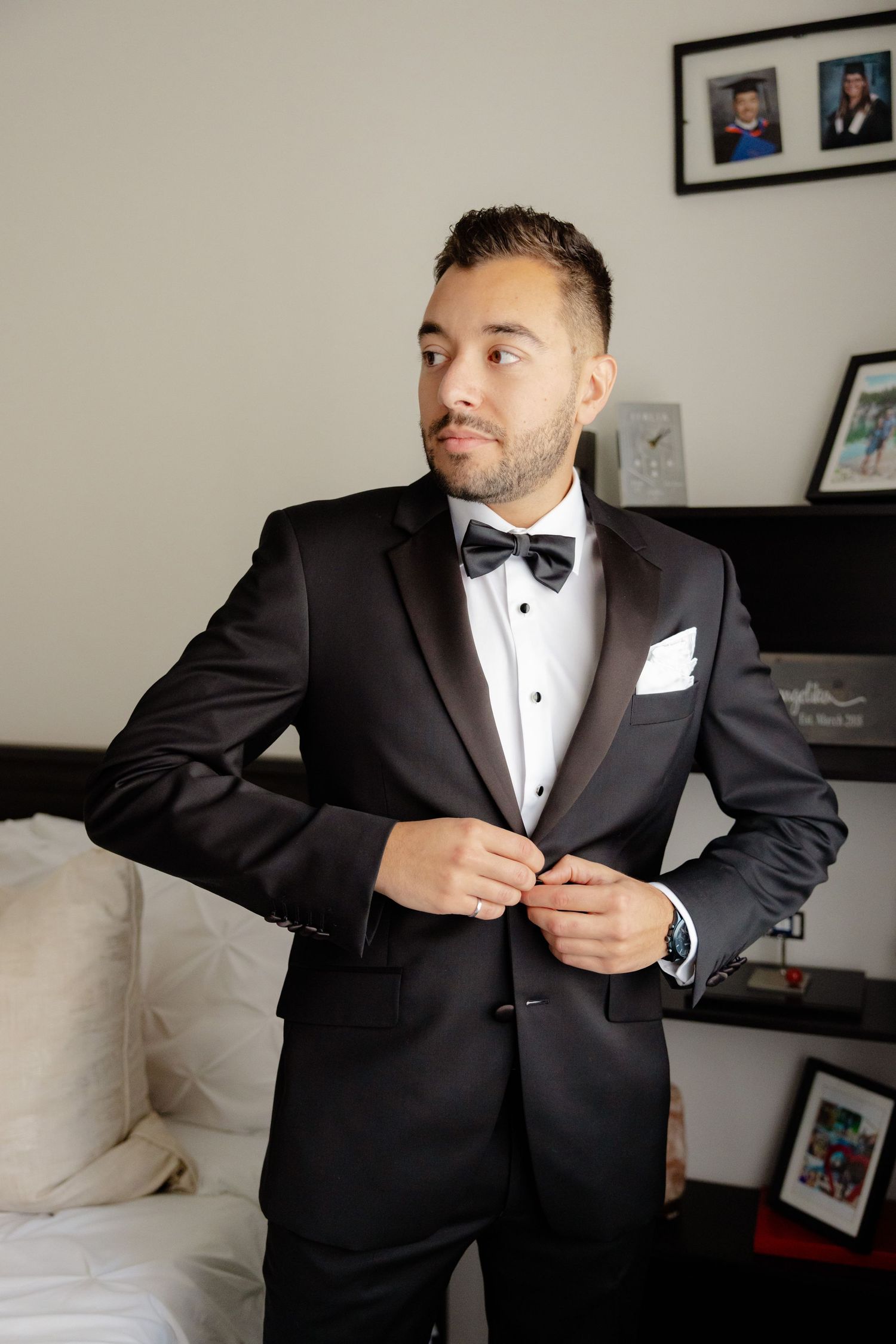 Person in black tuxedo and bow tie adjusts jacket while standing in bedroom.