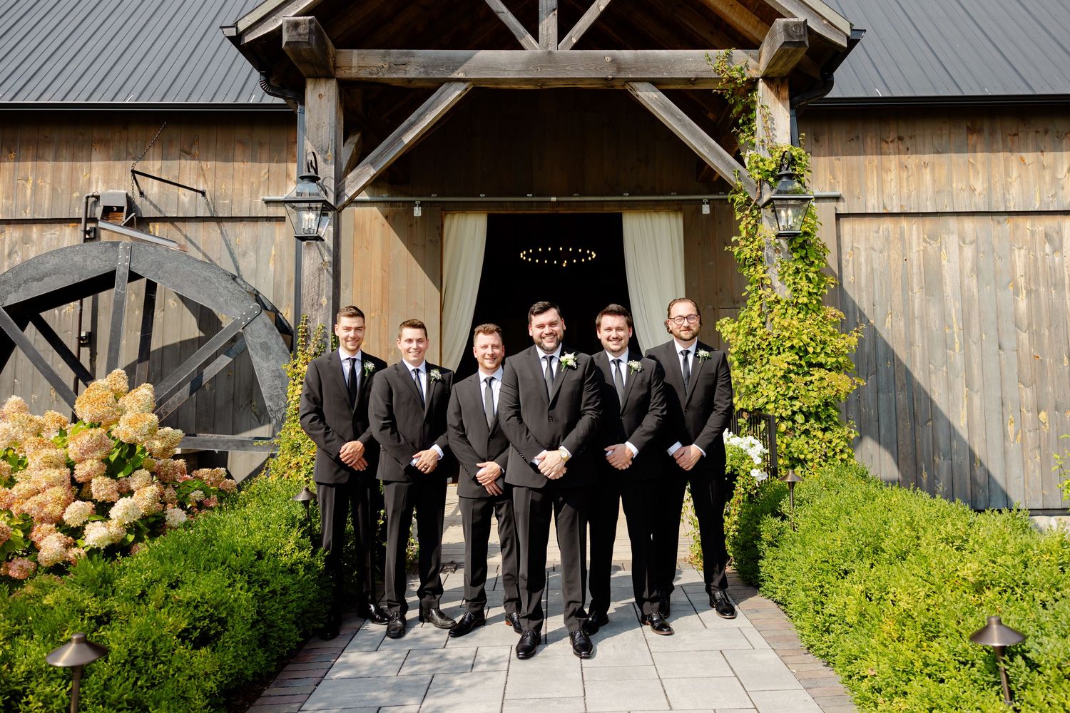 Formal group poses in matching dark suits outside a rustic barn entrance with landscaped path.