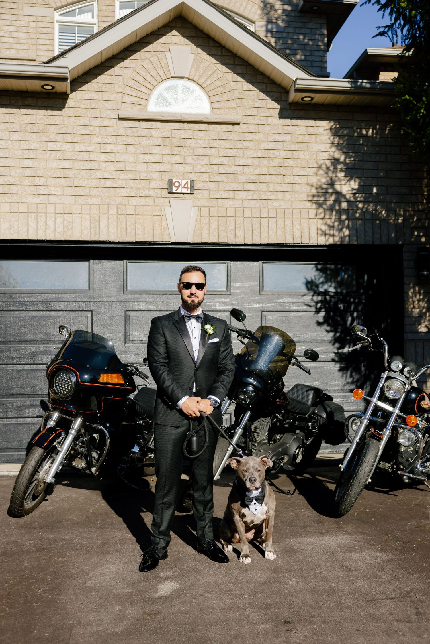 A formally dressed person stands with a dog between two motorcycles outside a garage.