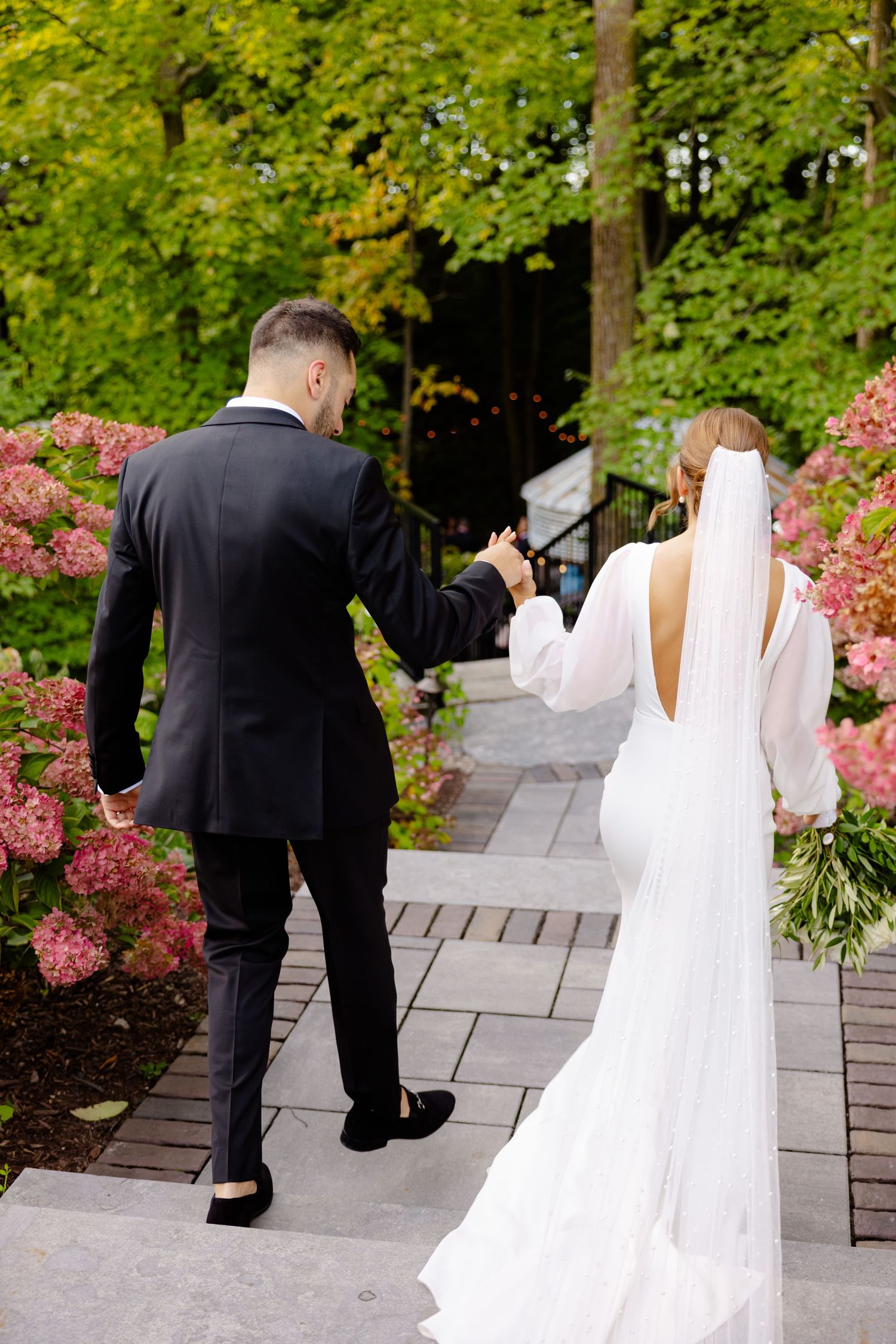 A couple walks hand in hand down a stone pathway surrounded by pink hydrangeas.