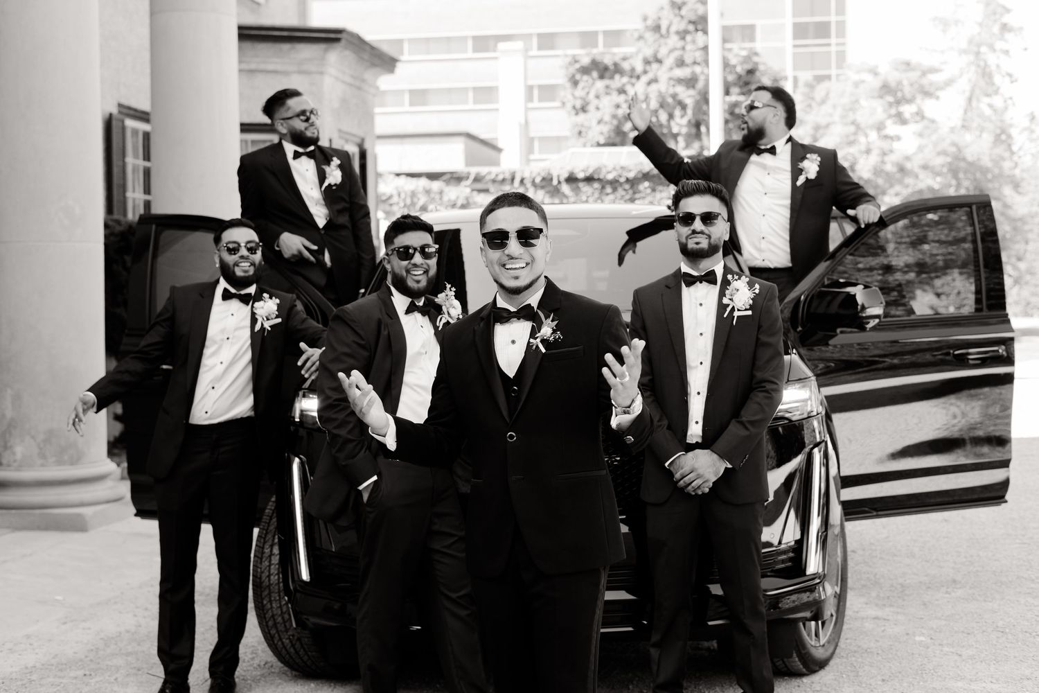 Wedding party members in tuxedos strike casual poses around a black vehicle at a formal venue.