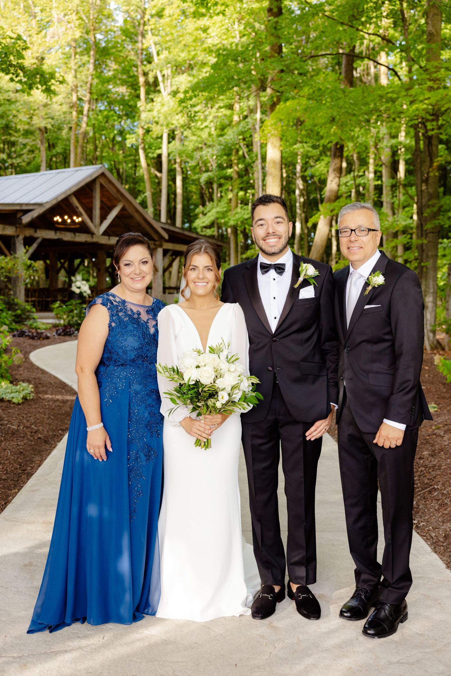 Wedding party in formal attire poses on concrete path with rustic wooden pavilion and forest backdrop.