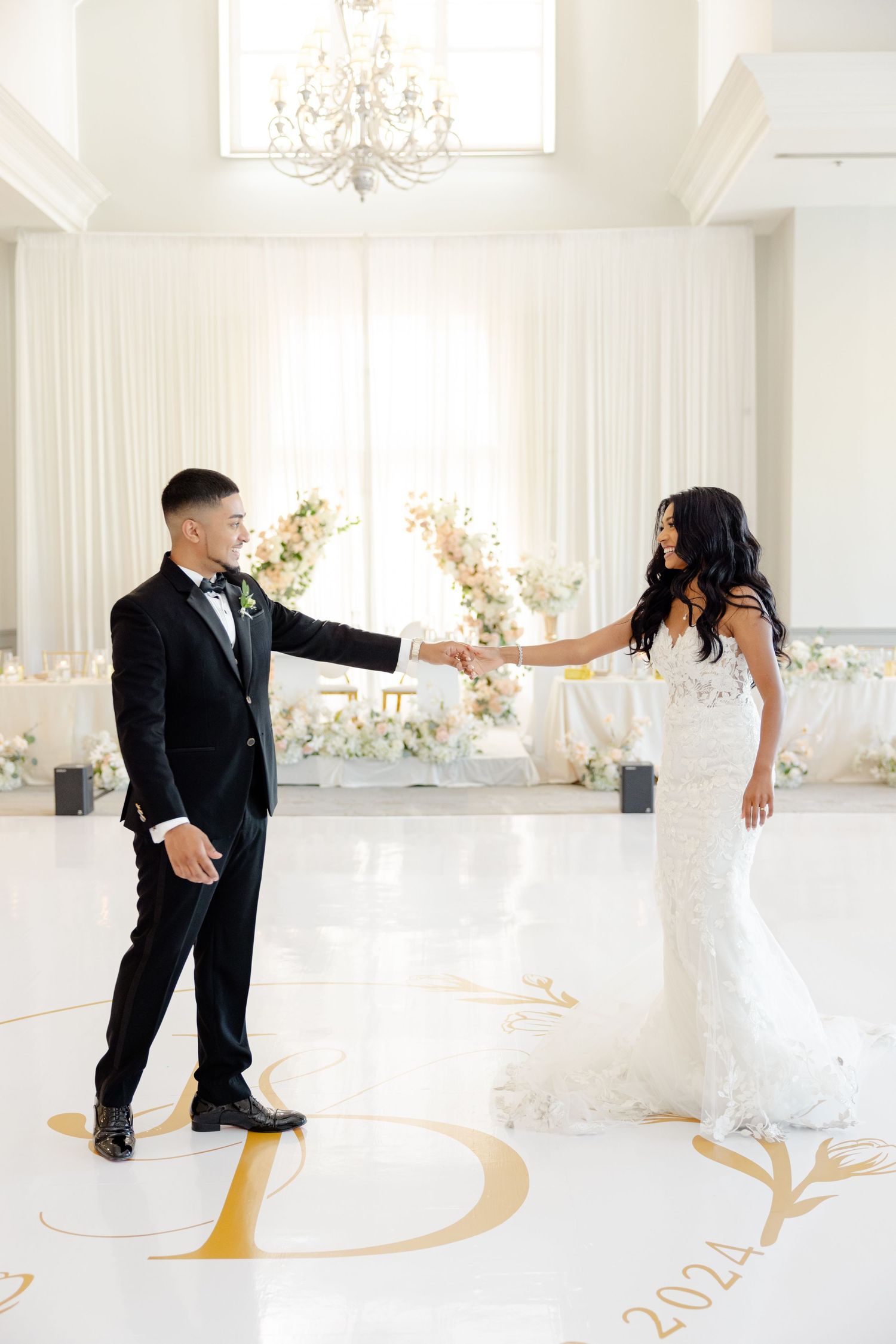 Dancing couple holds hands in bright wedding venue decorated with white draping and gold floor monogram.