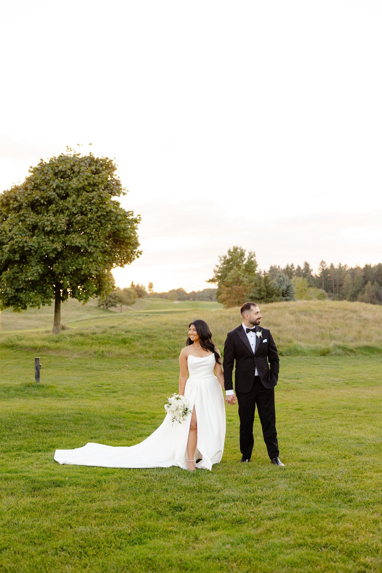 A wedding couple poses in an elegant outdoor setting with a flowing white dress and black tuxedo on a grassy hill.