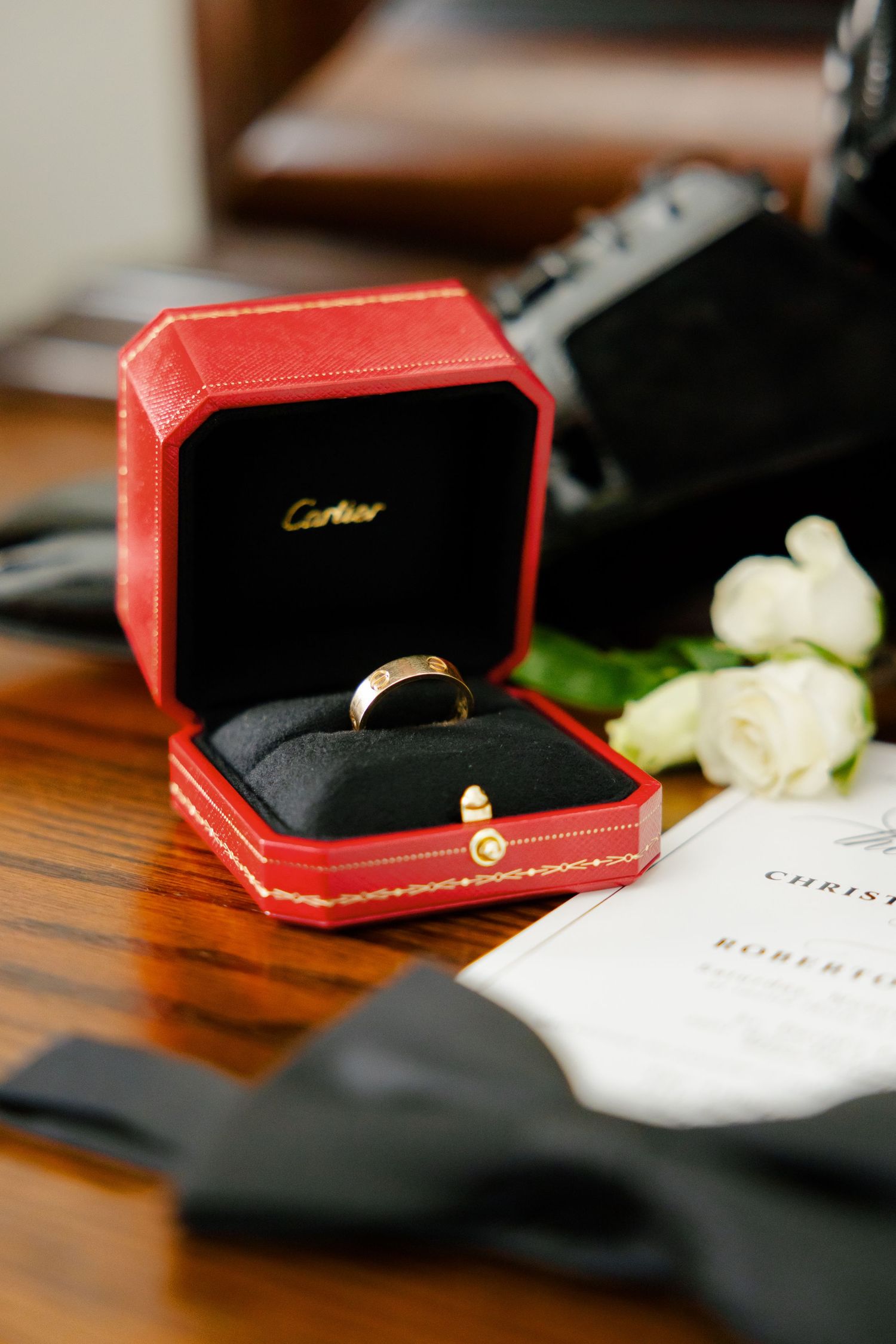 Cartier red ring box with gold band beside white roses on wooden table.