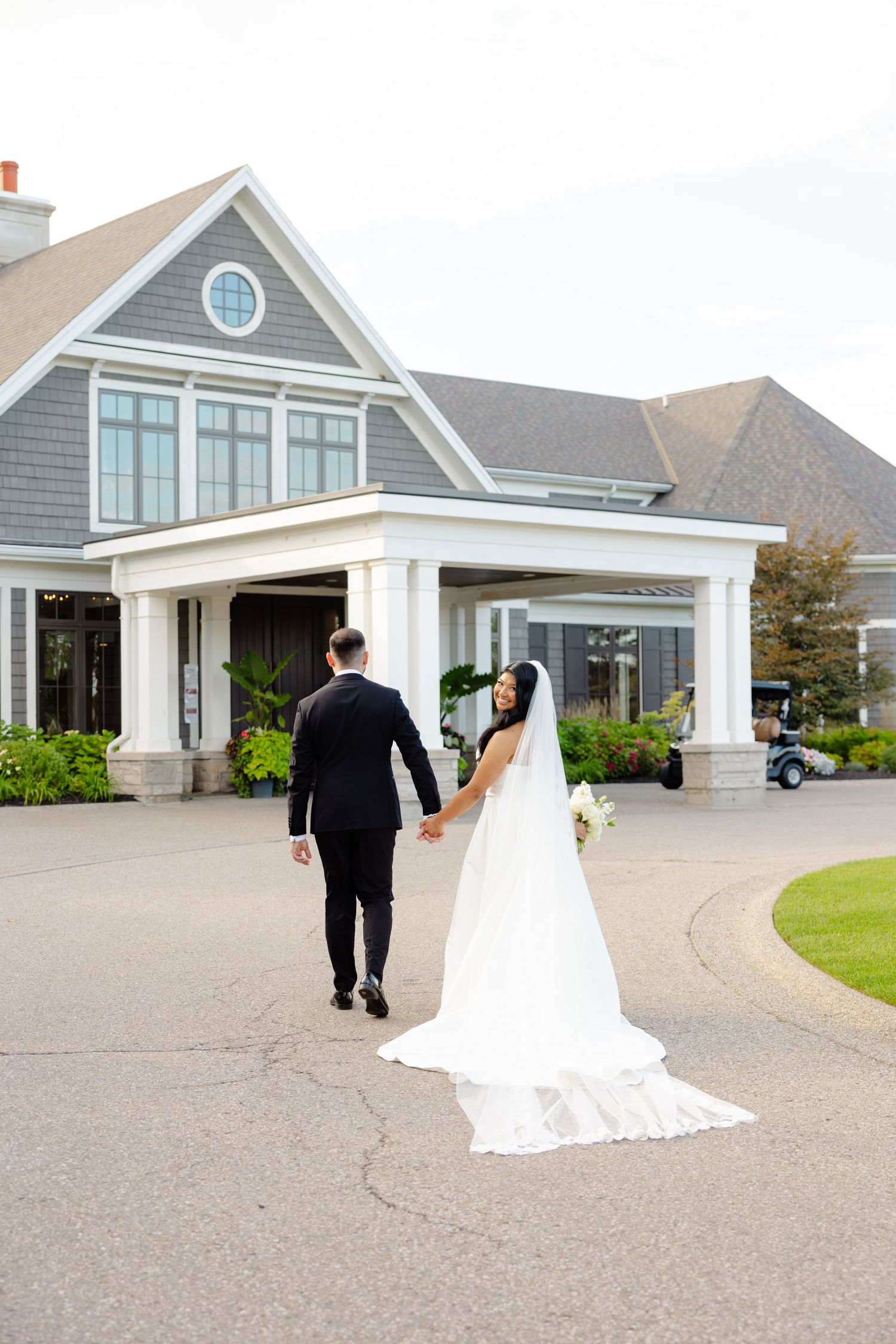A couple in wedding attire walks hand in hand toward a coastal-style gray house with white trim.