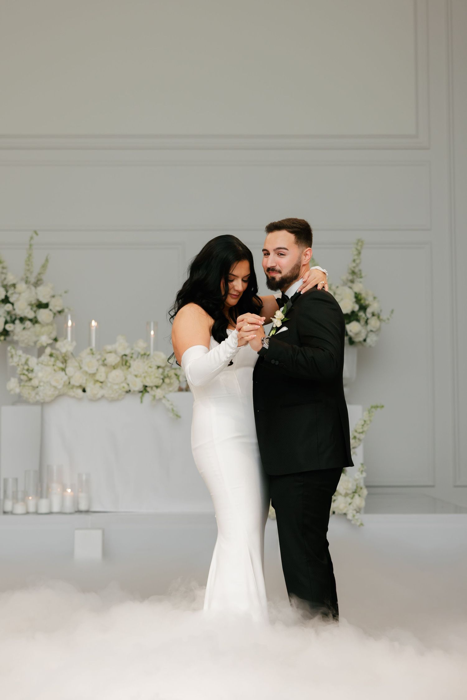 Couple shares intimate moment during first dance with white floral decor and fog effects.