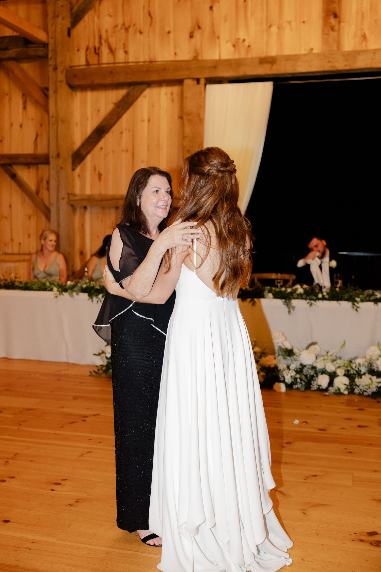 Two people share a dance in a rustic barn venue with decorated tables and floral arrangements visible.