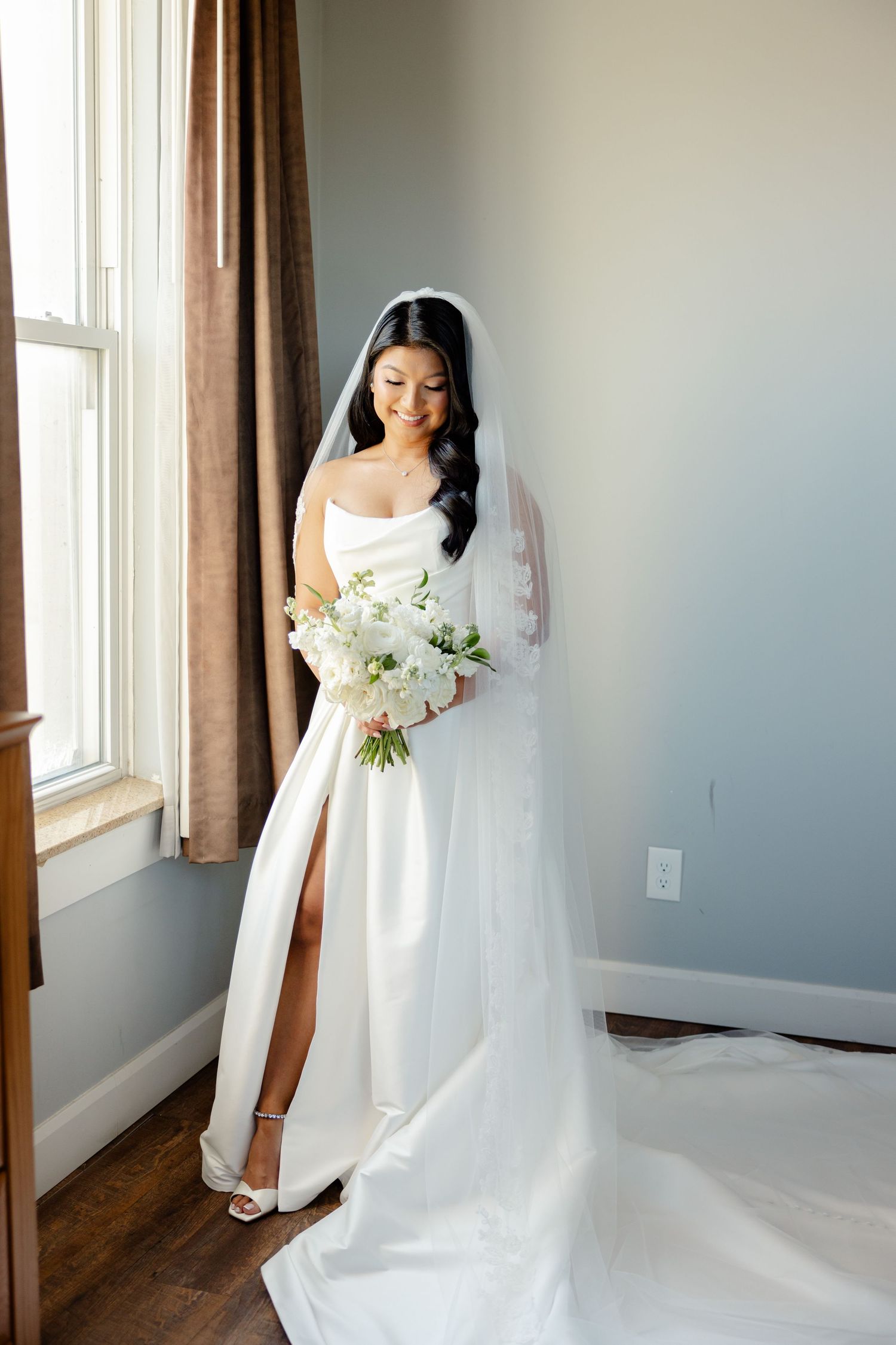 Bride in sleek white gown holds classic white bouquet while standing near window with natural light.