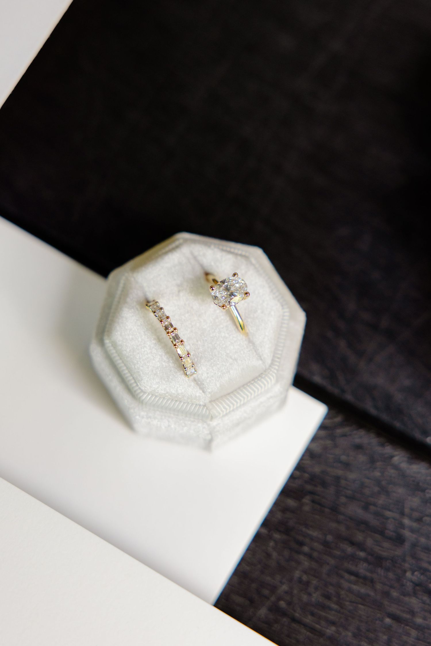 Delicate engagement and wedding rings displayed in white octagonal ring box against dark background.
