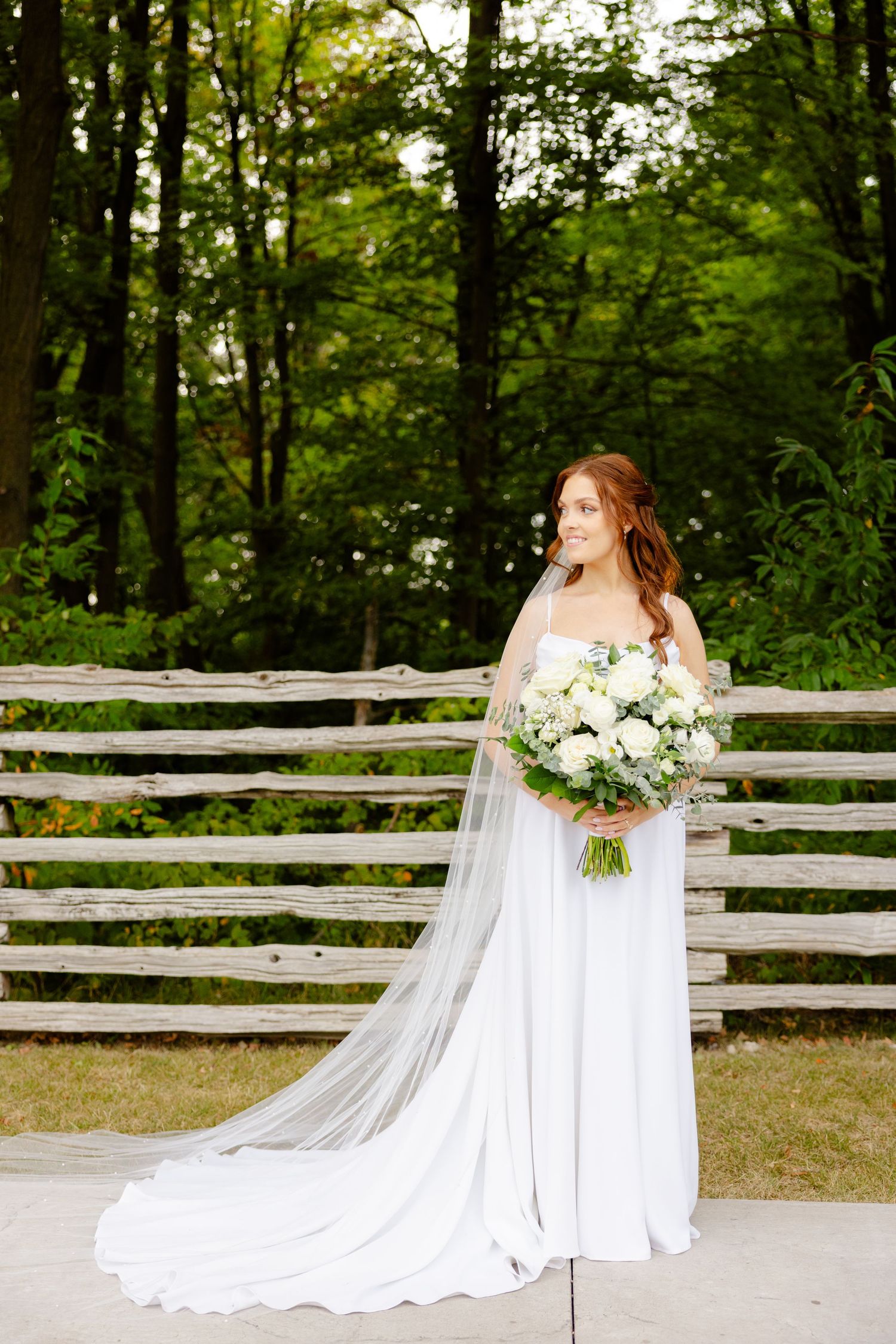 Bride in flowing white dress holds stunning bouquet against rustic outdoor backdrop.