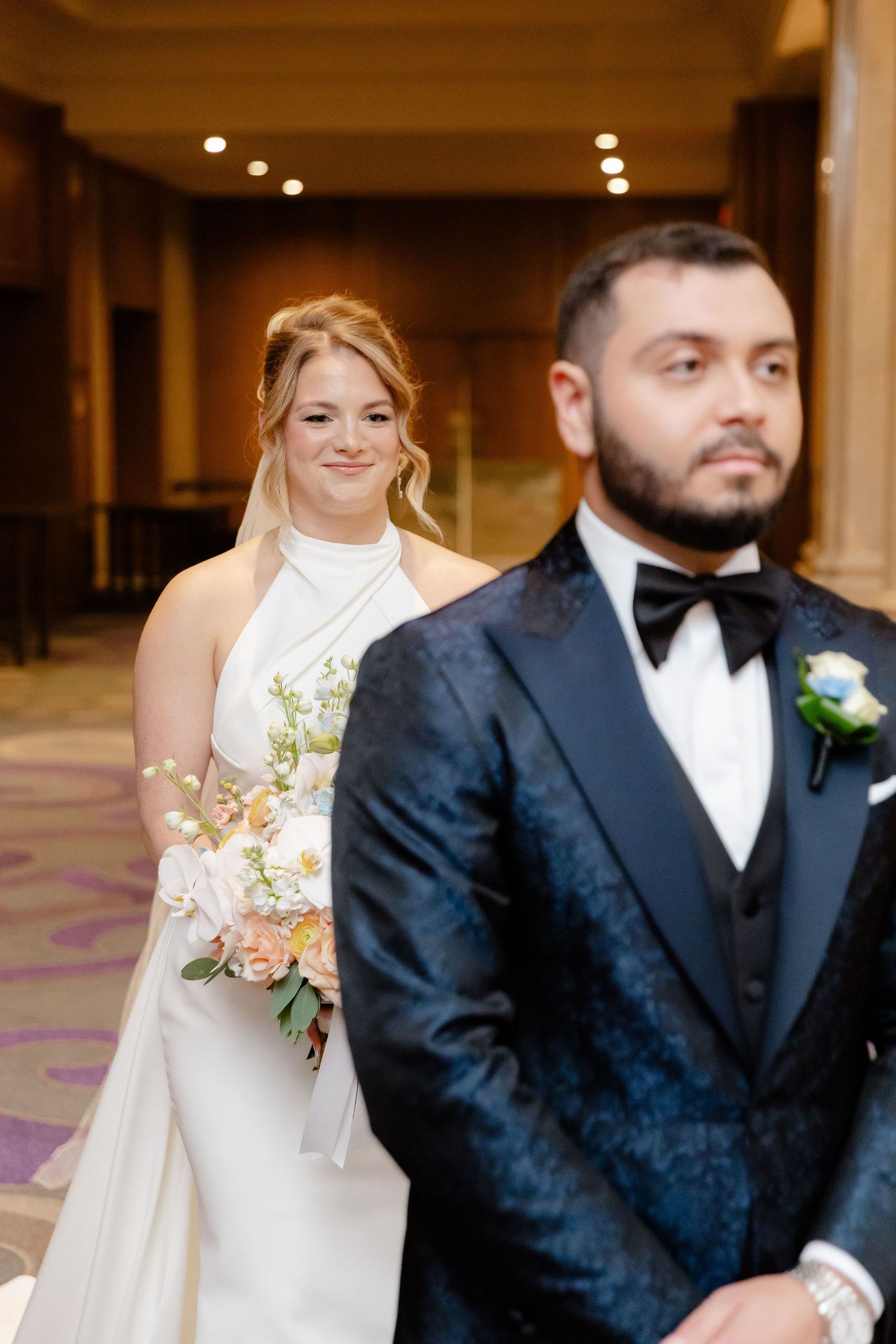A wedding couple poses in a hallway with elegant lighting, the bride in white and groom in navy tuxedo.