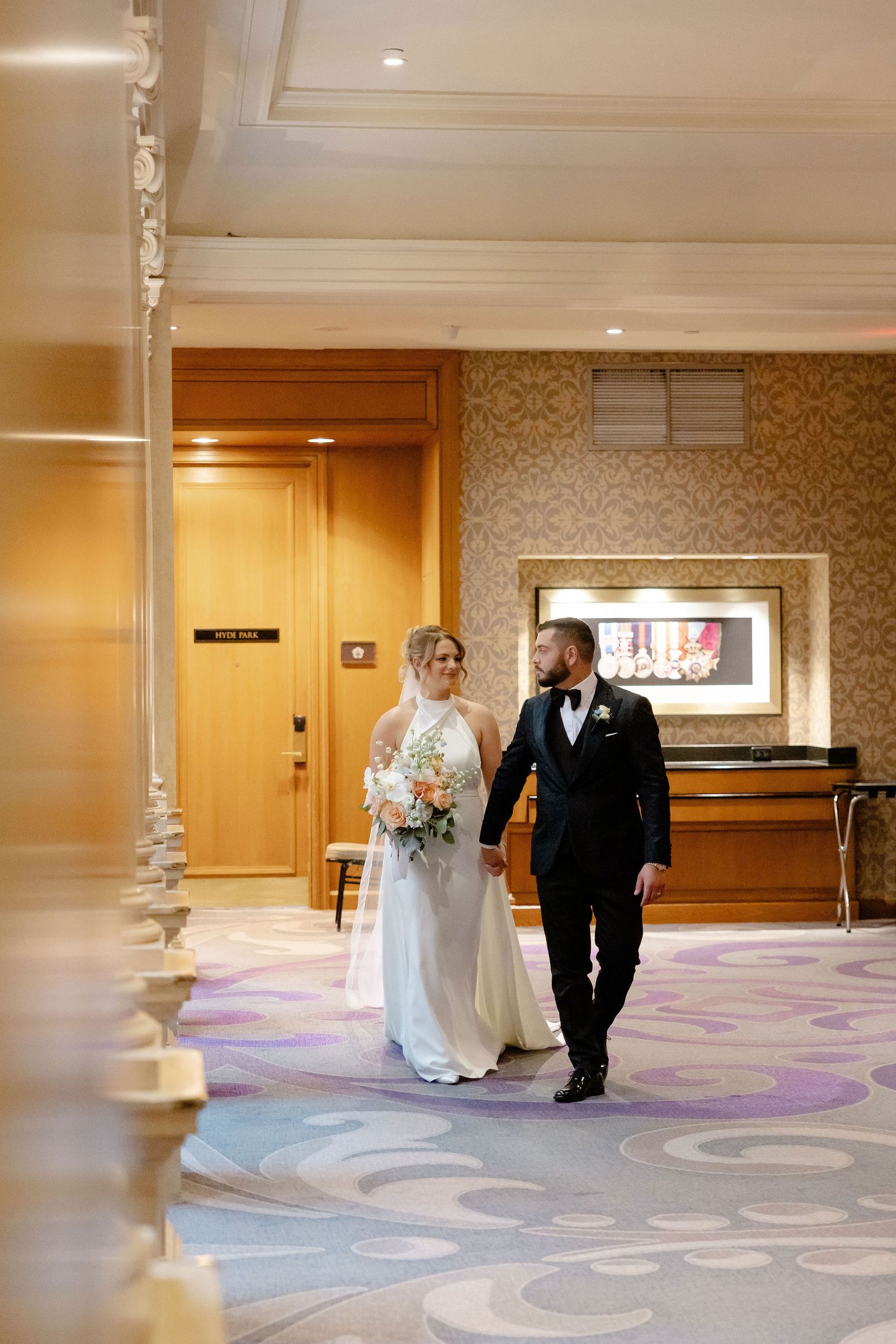 Wedding couple walks together through an elegant hotel hallway with patterned carpet and wood paneling.