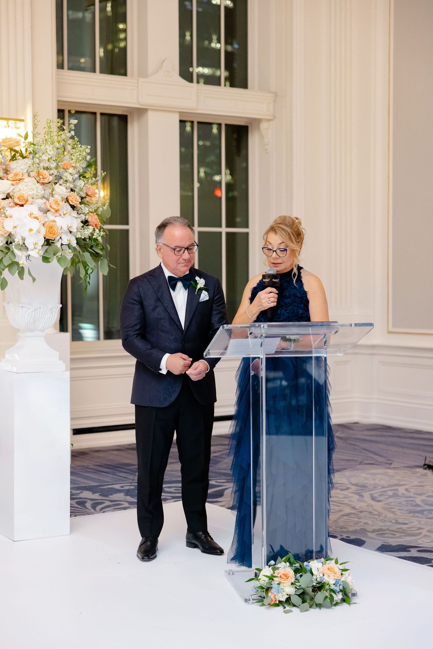 Wedding ceremony with a clear acrylic podium and white floral arrangements in an elegant white room.
