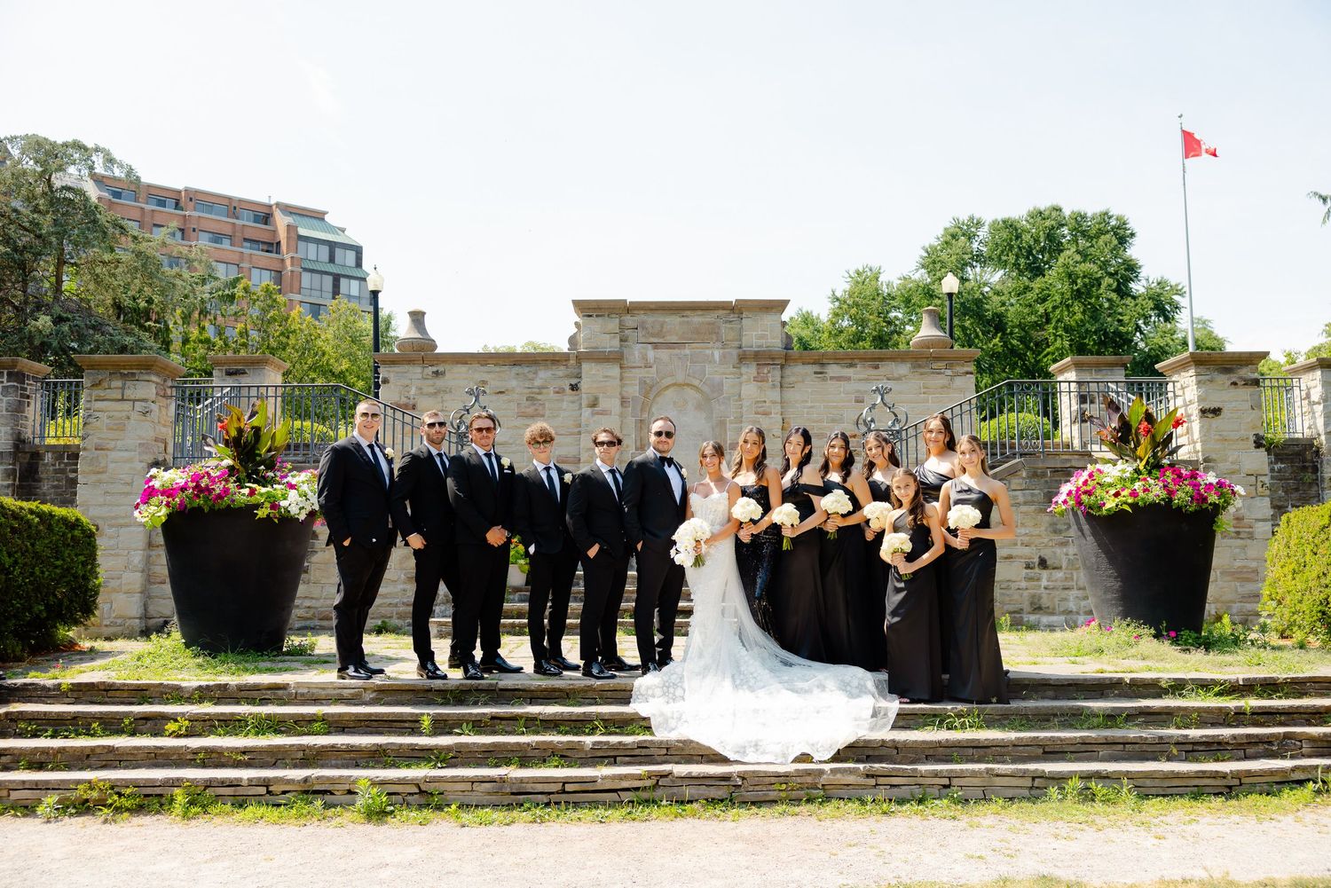 Formal wedding party arranged in a line poses outdoors on historic stone grounds with scenic greenery in the background.