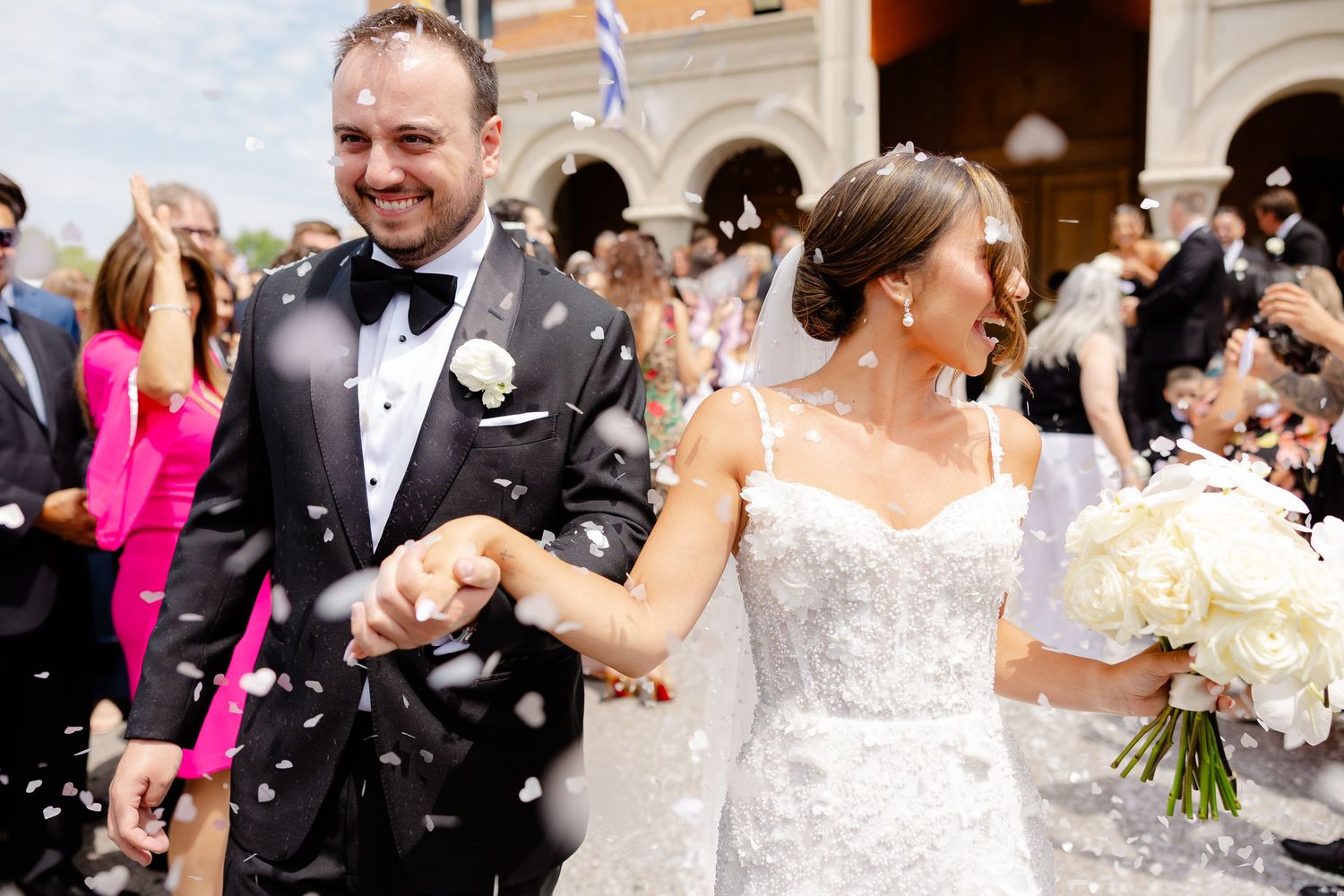 Wedding guests toss and celebrate with white flower petals during a joyous outdoor reception moment.