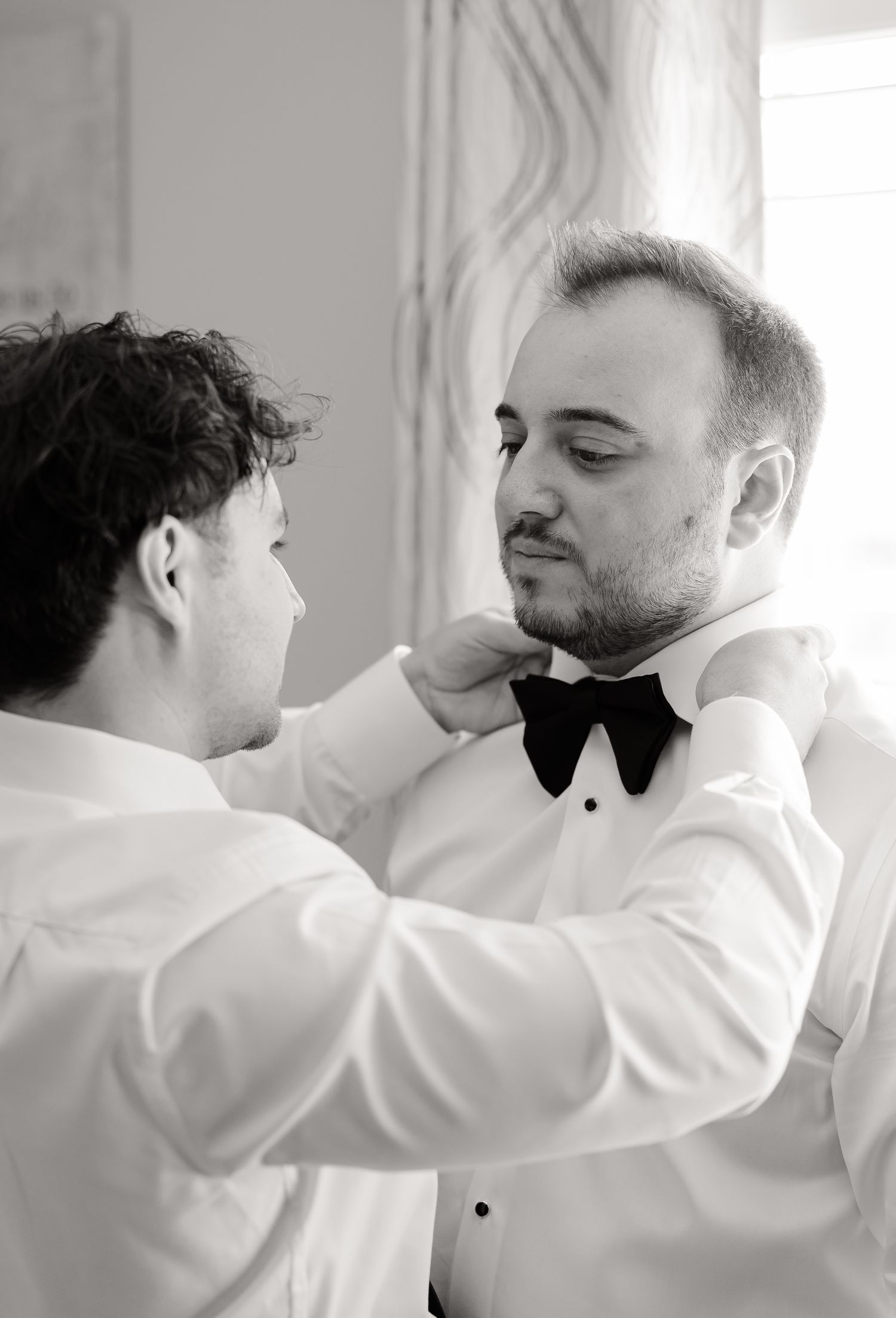 Someone helps adjust a black bow tie on a groom wearing a white tuxedo shirt in this elegant black and white photo.
