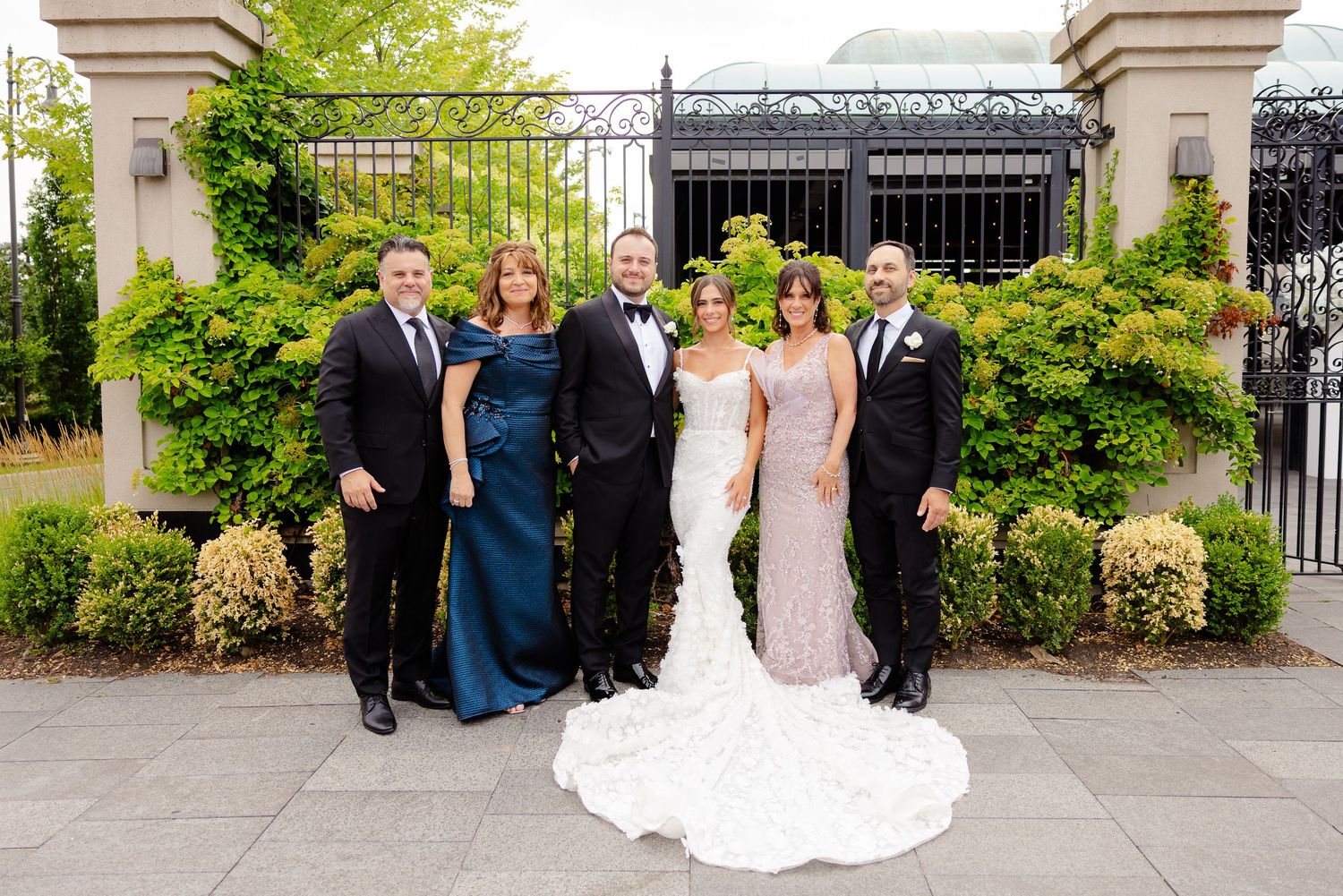 Elegant group in formal attire poses for wedding photo in front of garden gate with lush greenery and stone pillars.