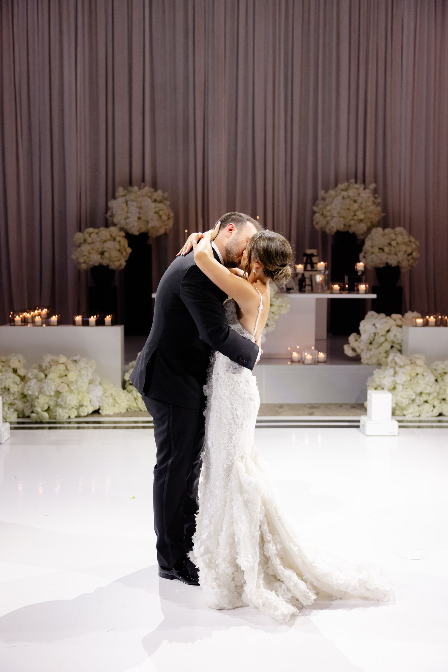 Two newlyweds share their first dance on a white floor surrounded by elegant white floral arrangements and candlelight.