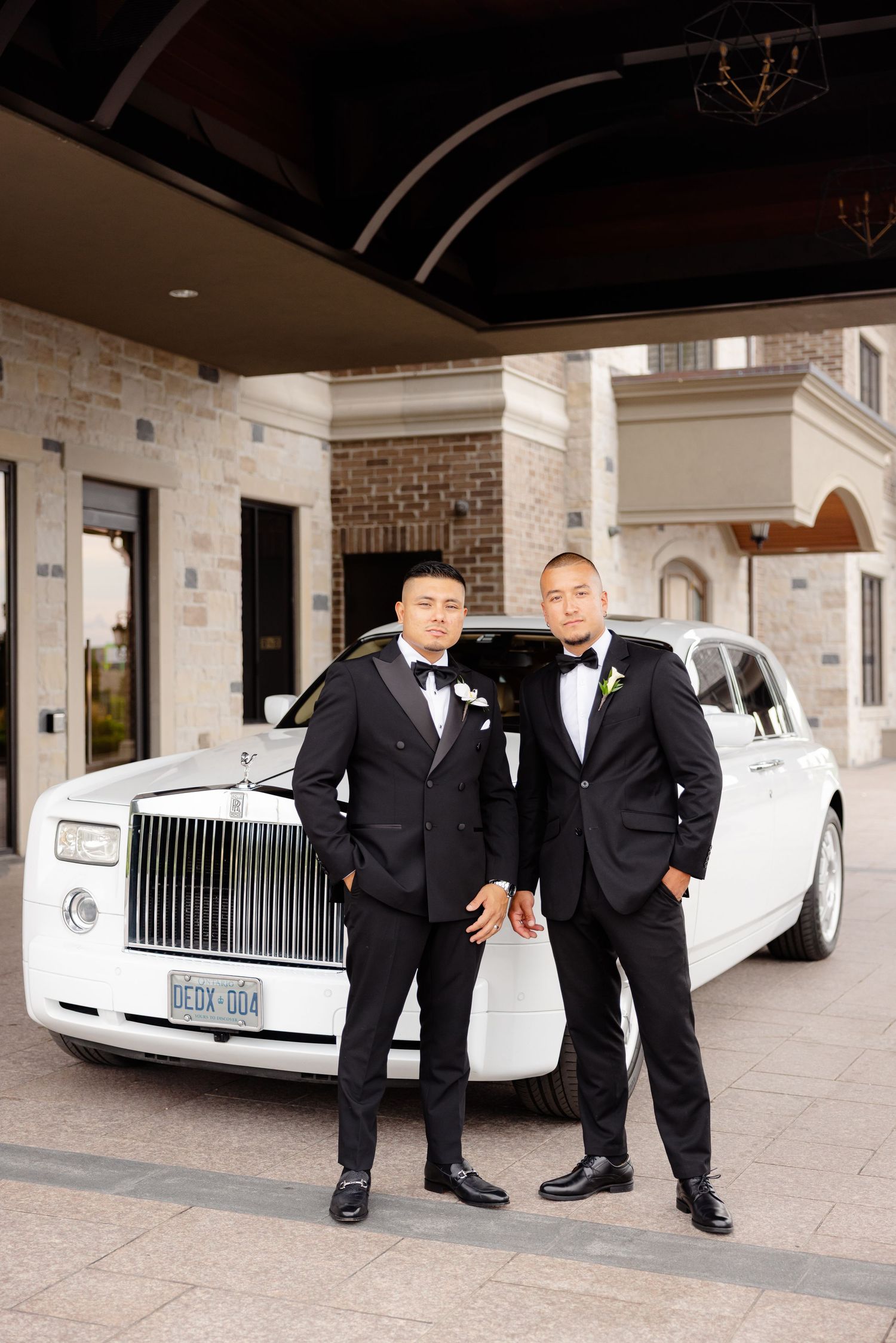 Groomsmen in black tuxedos pose alongside white Rolls-Royce luxury cars at a brick building venue entrance.