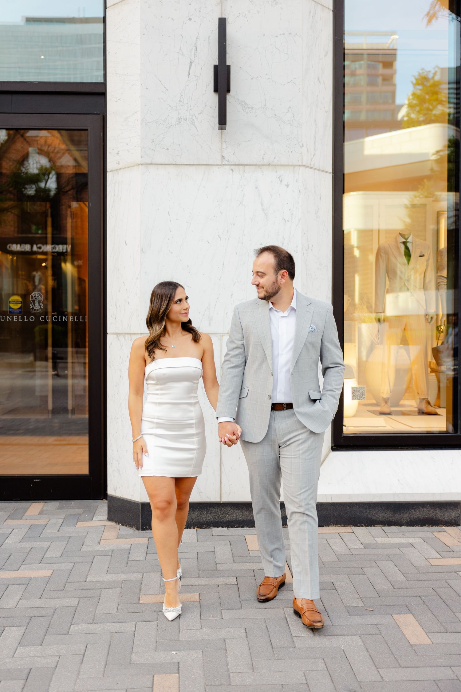 Couple walks hand in hand along urban sidewalk wearing coordinated formal outfits.