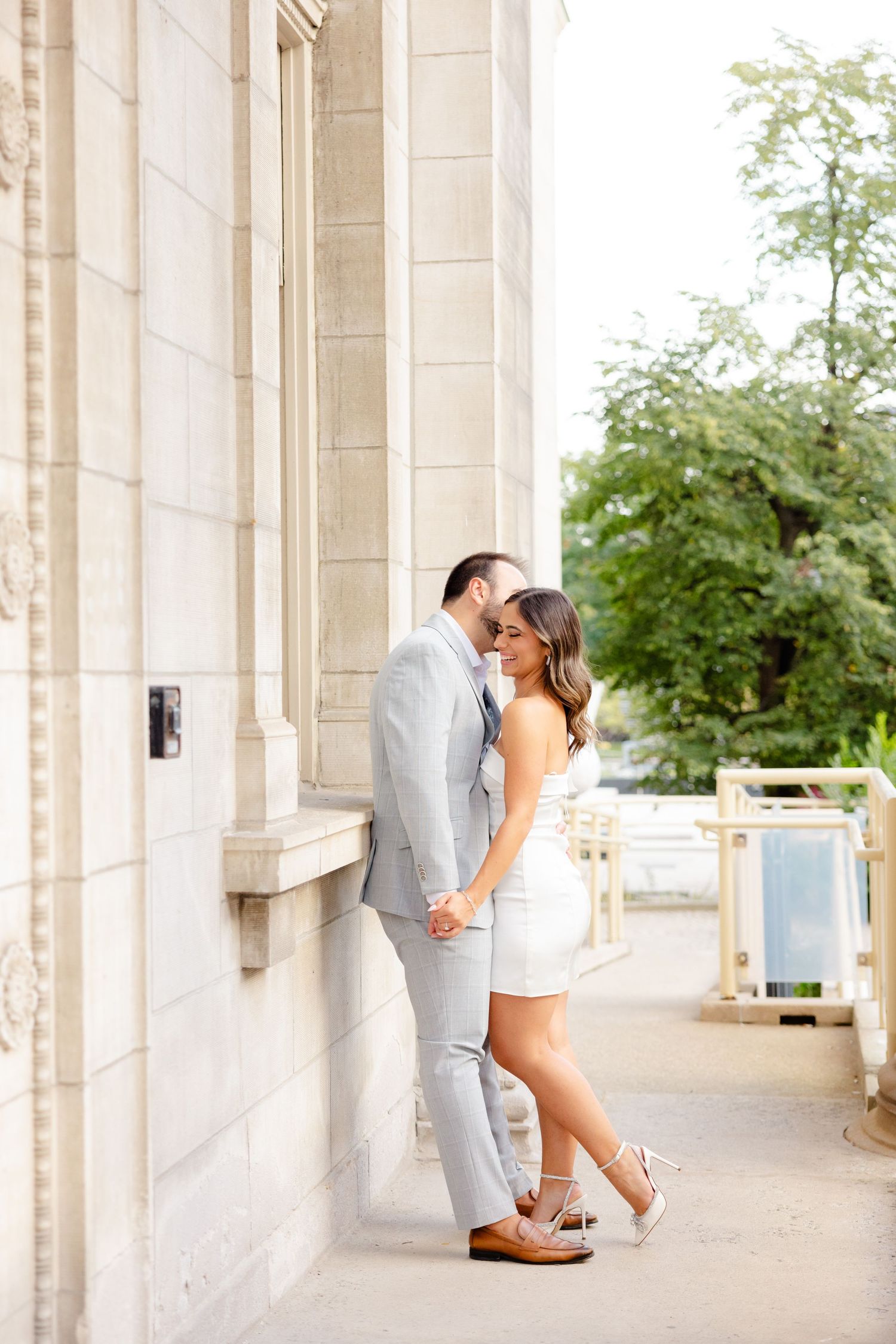 A couple shares a tender moment while standing against a stone pillar on a covered walkway.