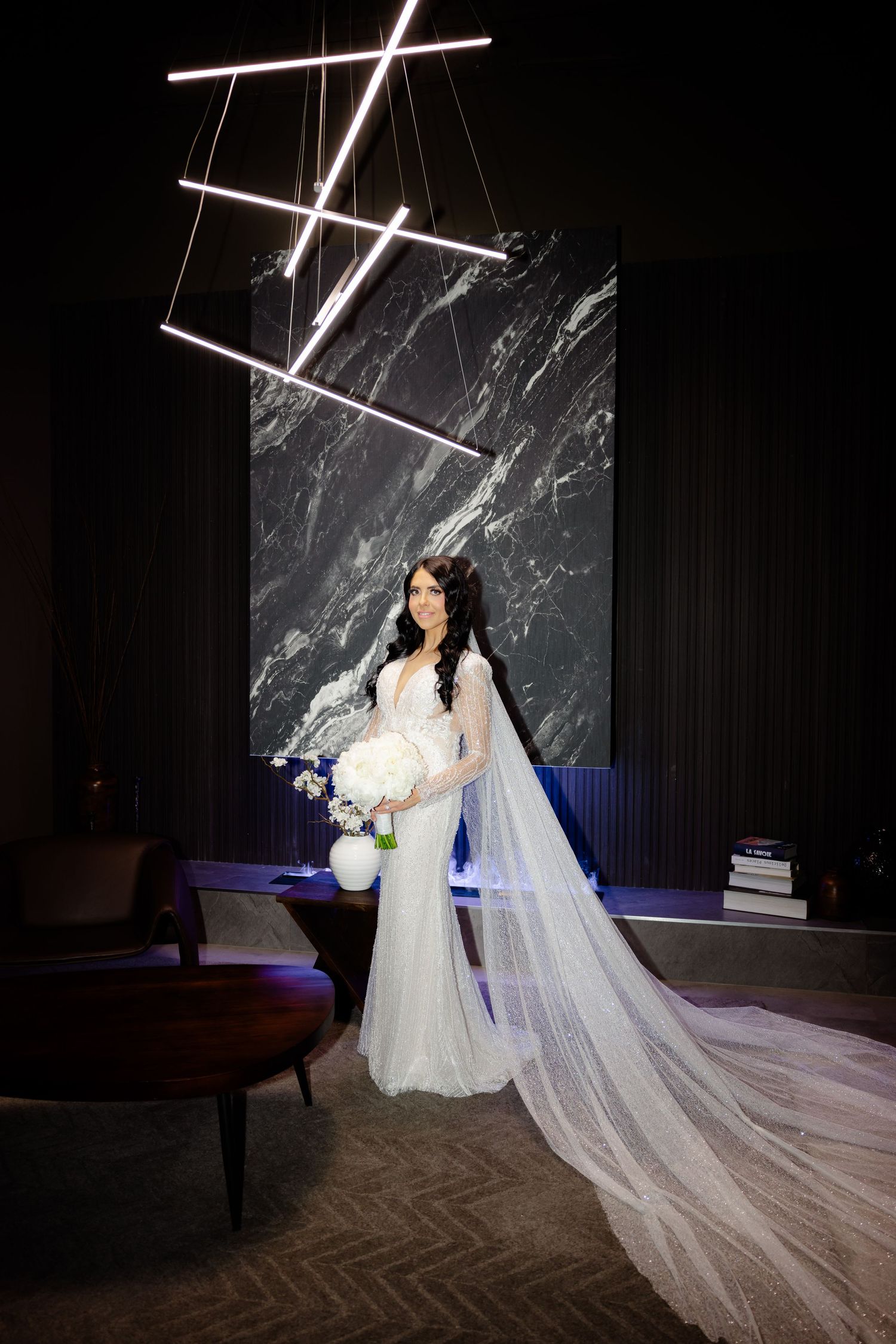 Elegant bride in a lace gown with cathedral veil poses against dark marble wall with modern lighting.