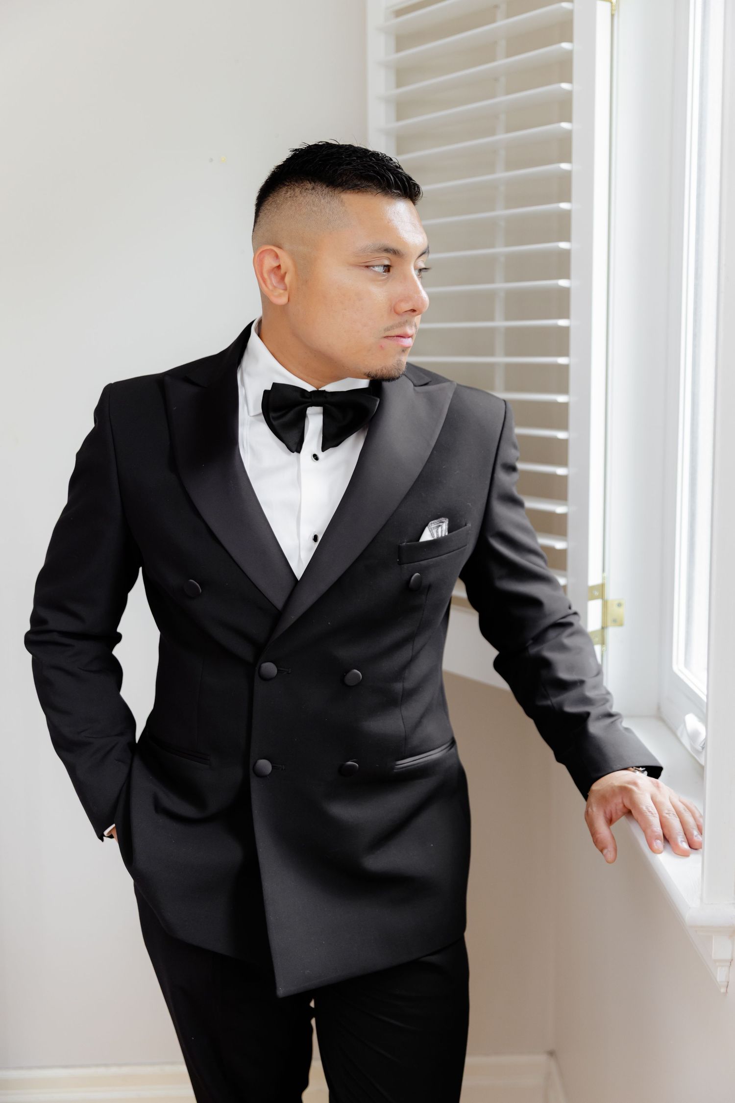 A groom in a classic black tuxedo peers through white window blinds during a formal wedding photoshoot.