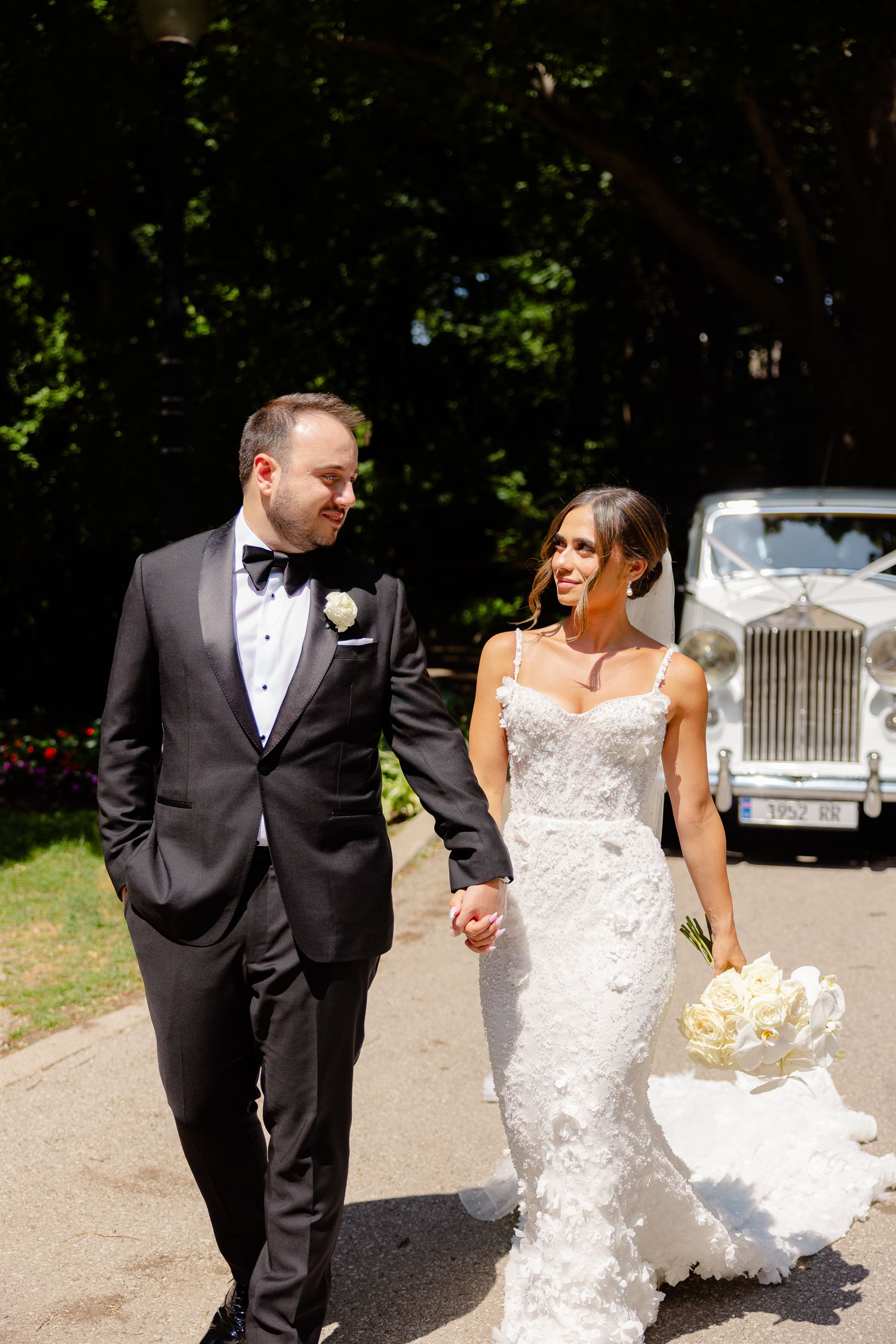 A couple in wedding attire walk hand in hand on a sunny path with a vintage Rolls Royce visible in the background.