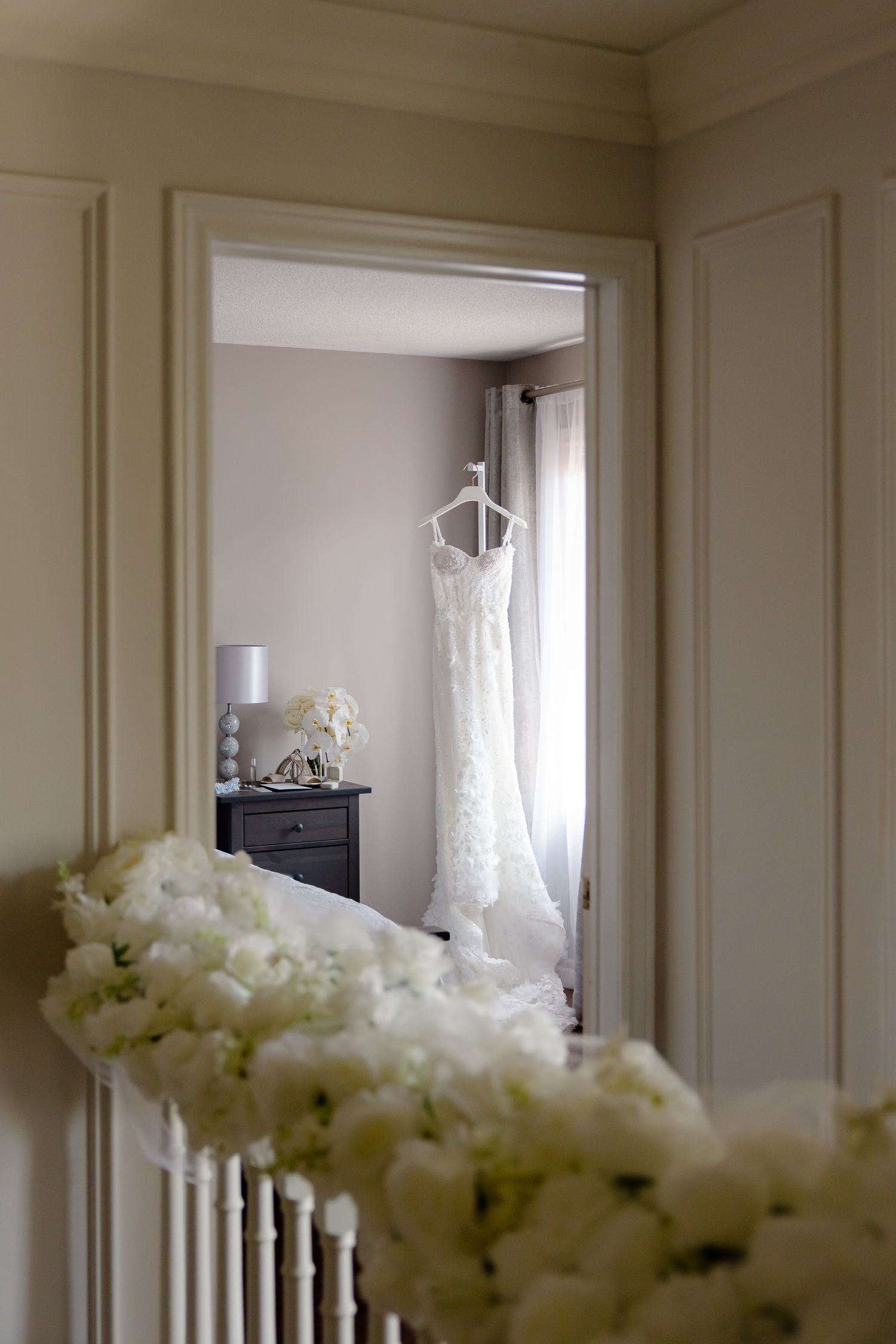 An elegant white lace wedding dress hangs in a doorway with white floral decorations in the foreground.