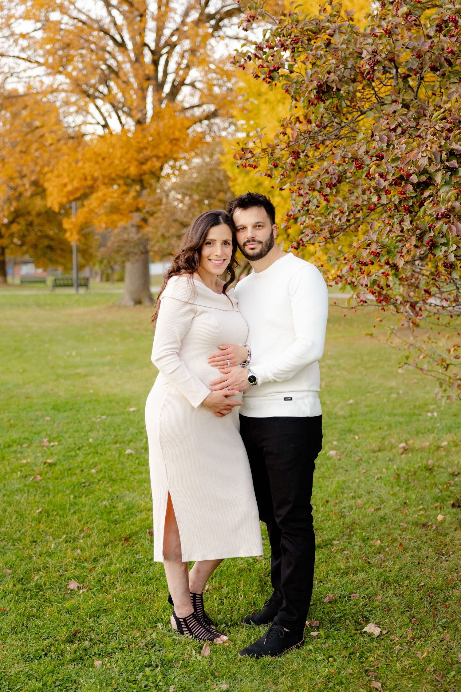 Couple in white outfits embracing in a park with autumn foliage and trees in the background.
