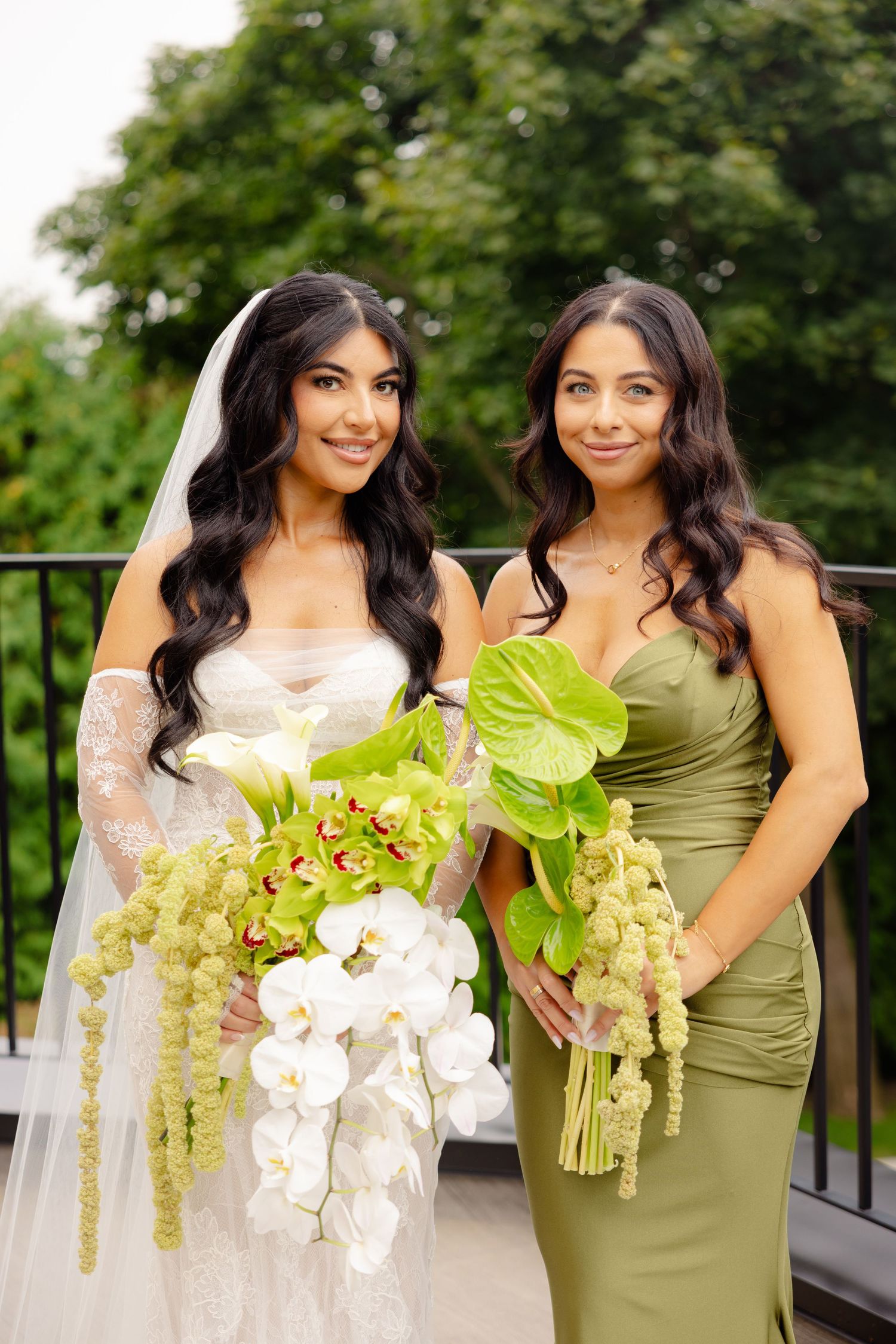 Two women in wedding party attire smile while holding elegant orchid bouquets against a lush outdoor backdrop.