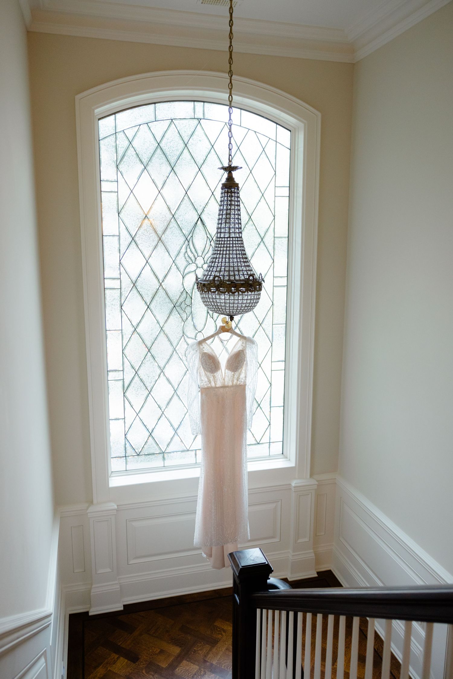 Elegant blush wedding dress hanging beneath crystal chandelier against diamond-paned window.