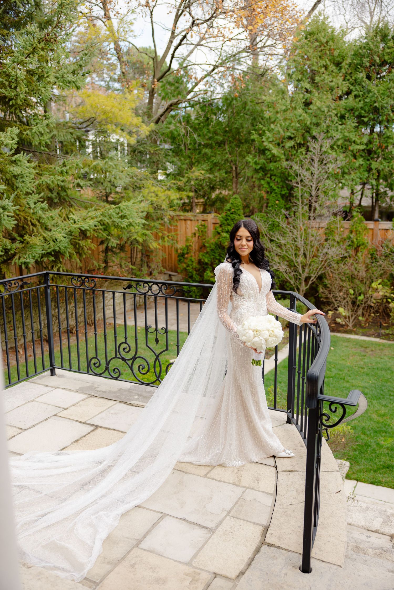 Bride in elegant fitted gown with cathedral length veil stands on stone terrace surrounded by autumn trees.