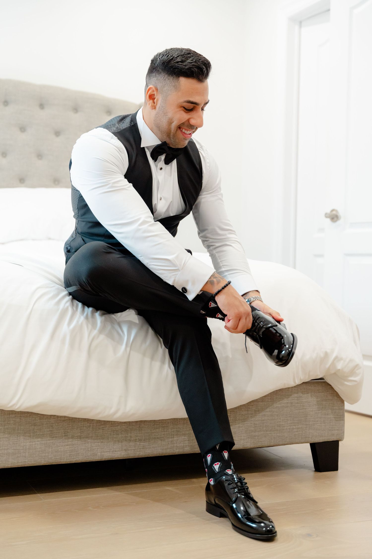 Person in formal attire adjusting dress shoes while sitting on white upholstered bed.