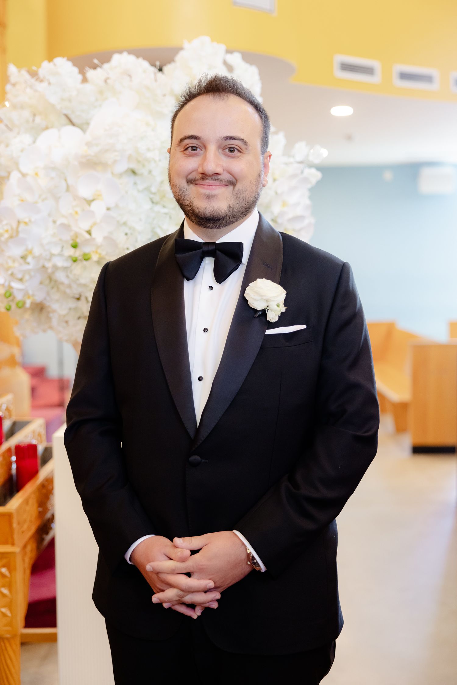 Wedding attendee in a classic black tuxedo with bow tie and white boutonniere poses in front of floral arrangements.