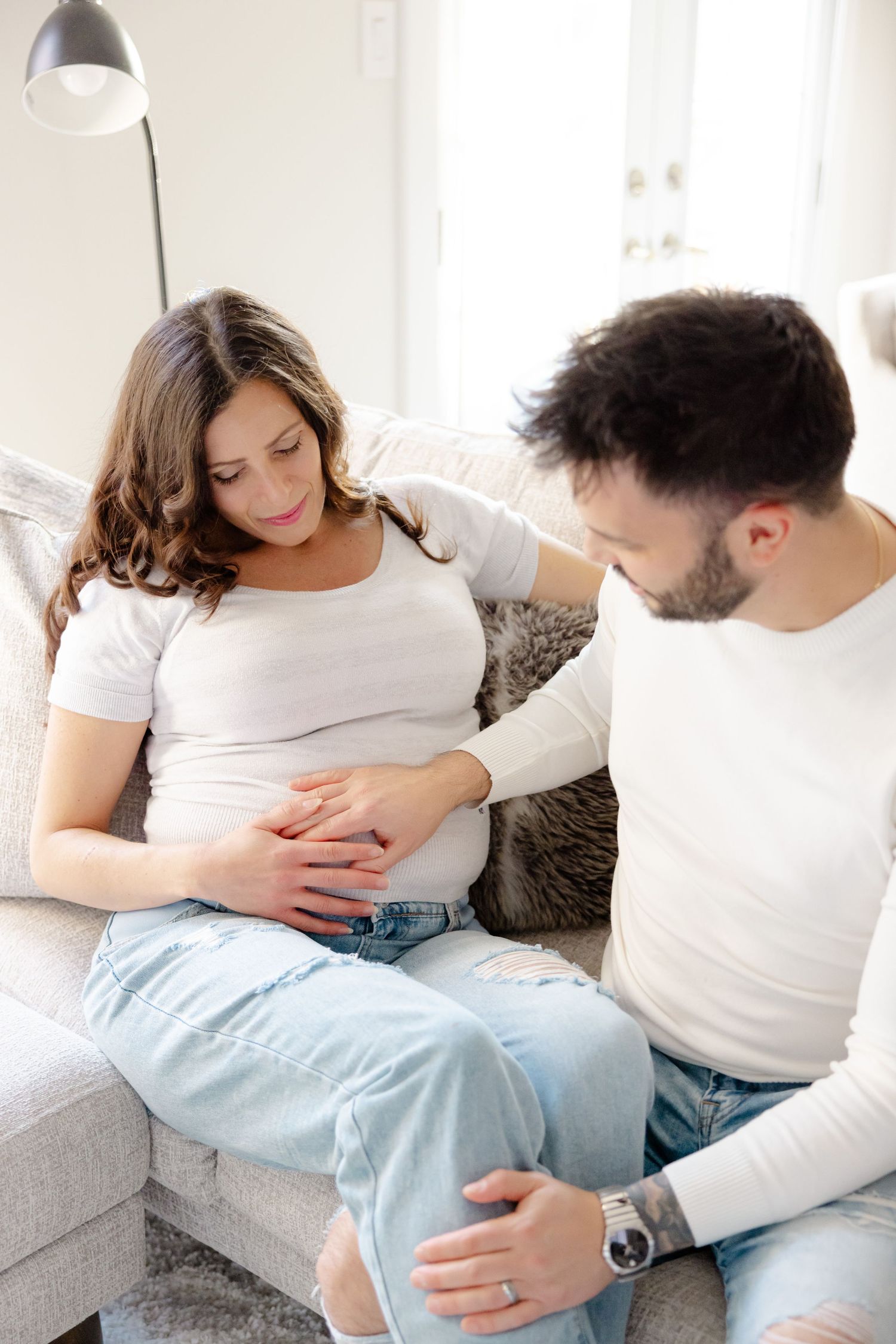 Couple sitting on couch, with the woman appearing pregnant and both touching her belly affectionately.
