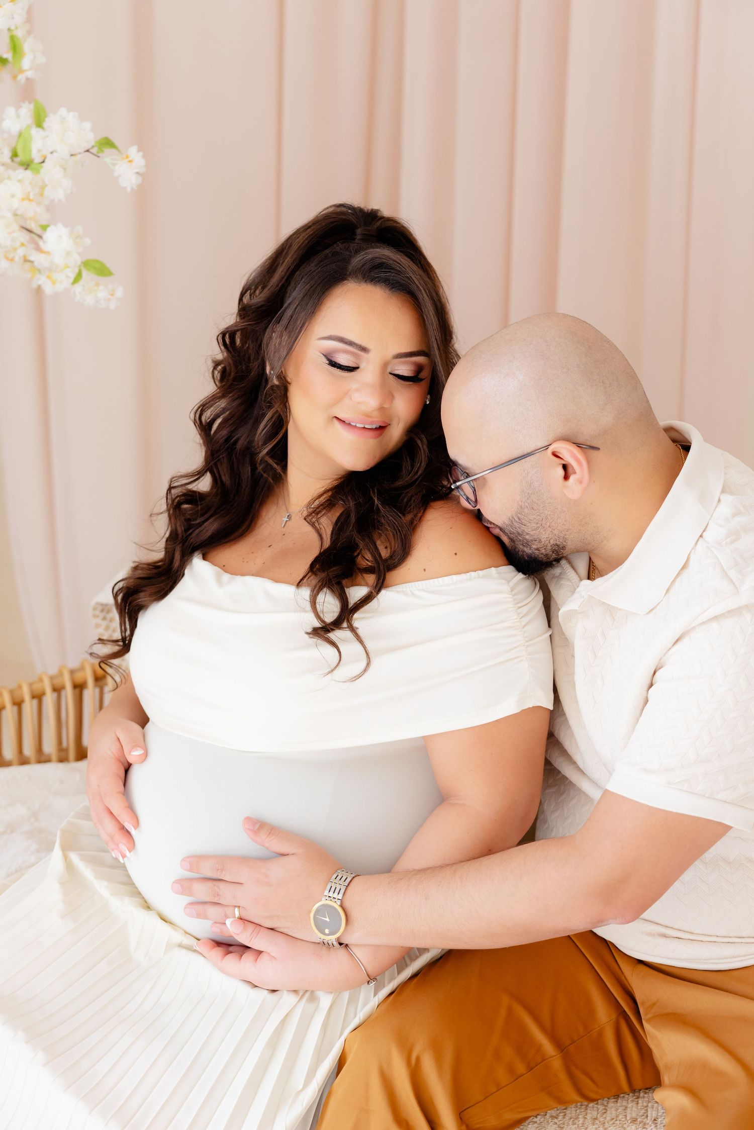 Intimate maternity photo featuring couple in white shirts against blush backdrop with white flowers.