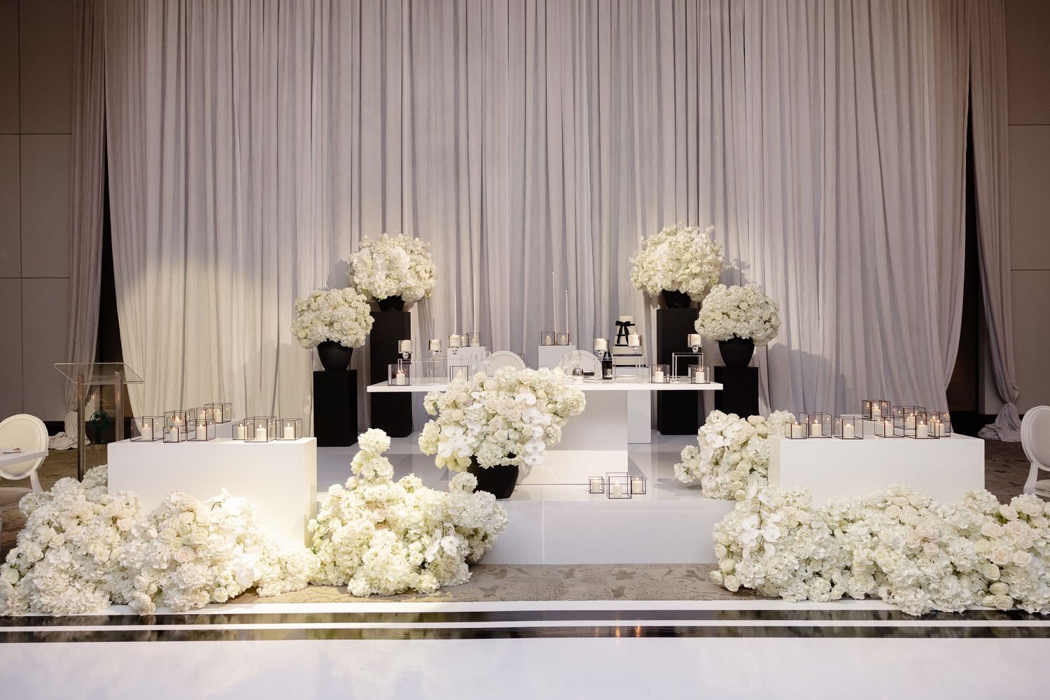 Elegant white wedding reception stage setup with cascading white flowers and black vases on tiered platforms.
