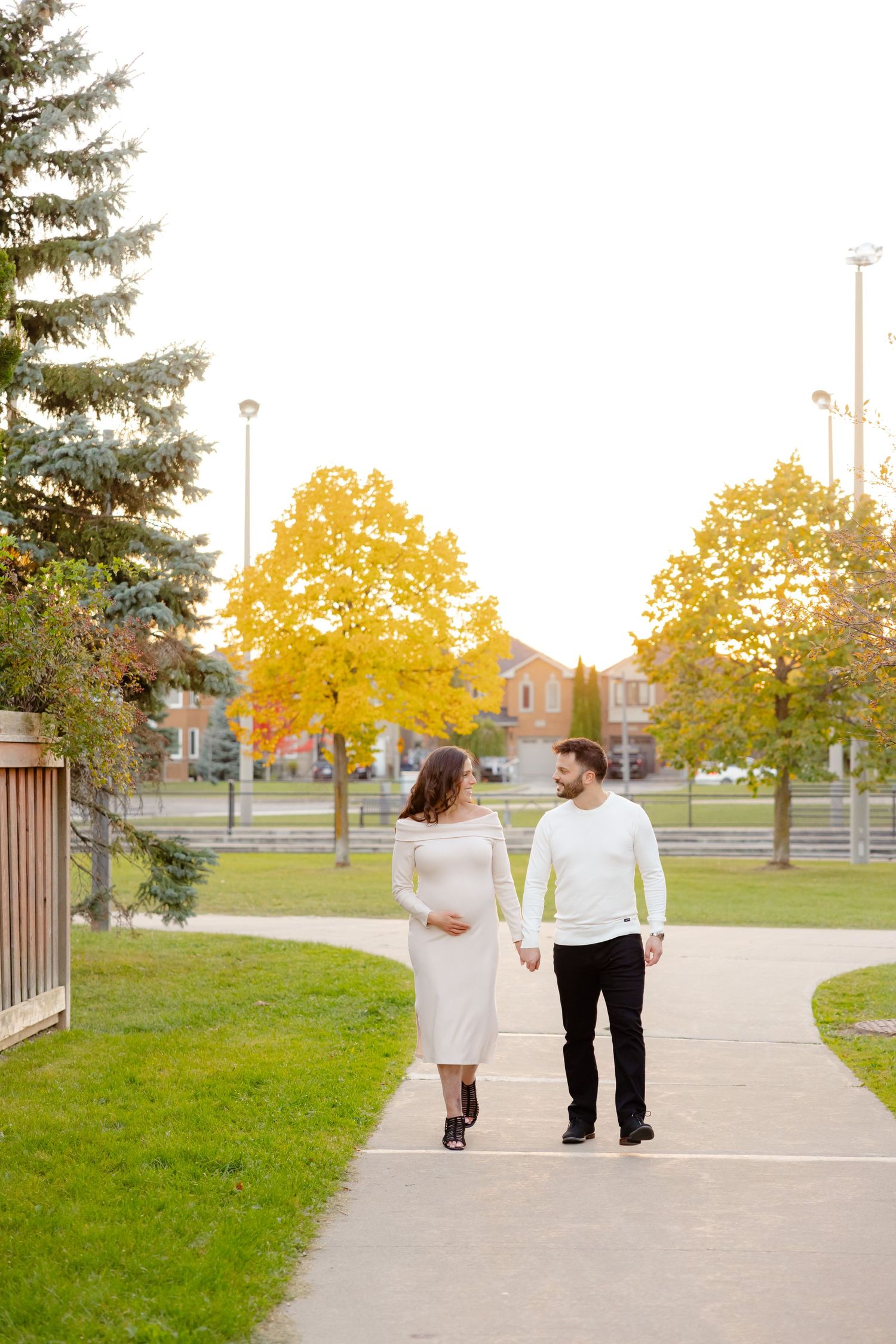 A couple walks hand in hand along a path surrounded by autumn trees in a suburban park setting.