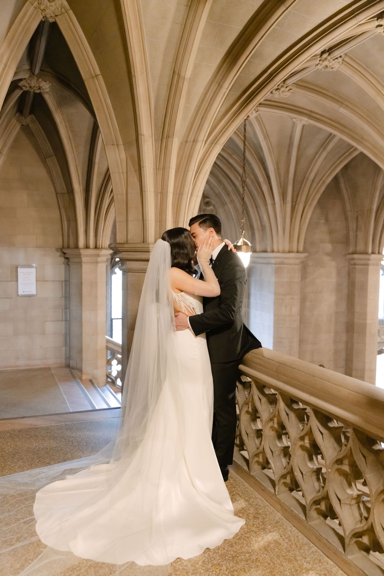 Wedding couple kiss on ornate stone staircase under vaulted cathedral ceiling.