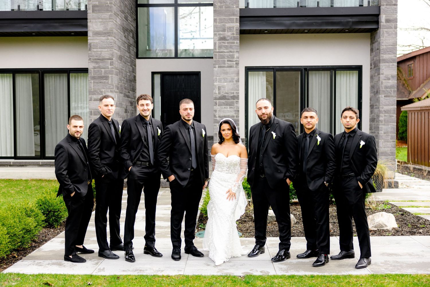 Wedding party in black suits poses with bride in white gown outside modern stone building.