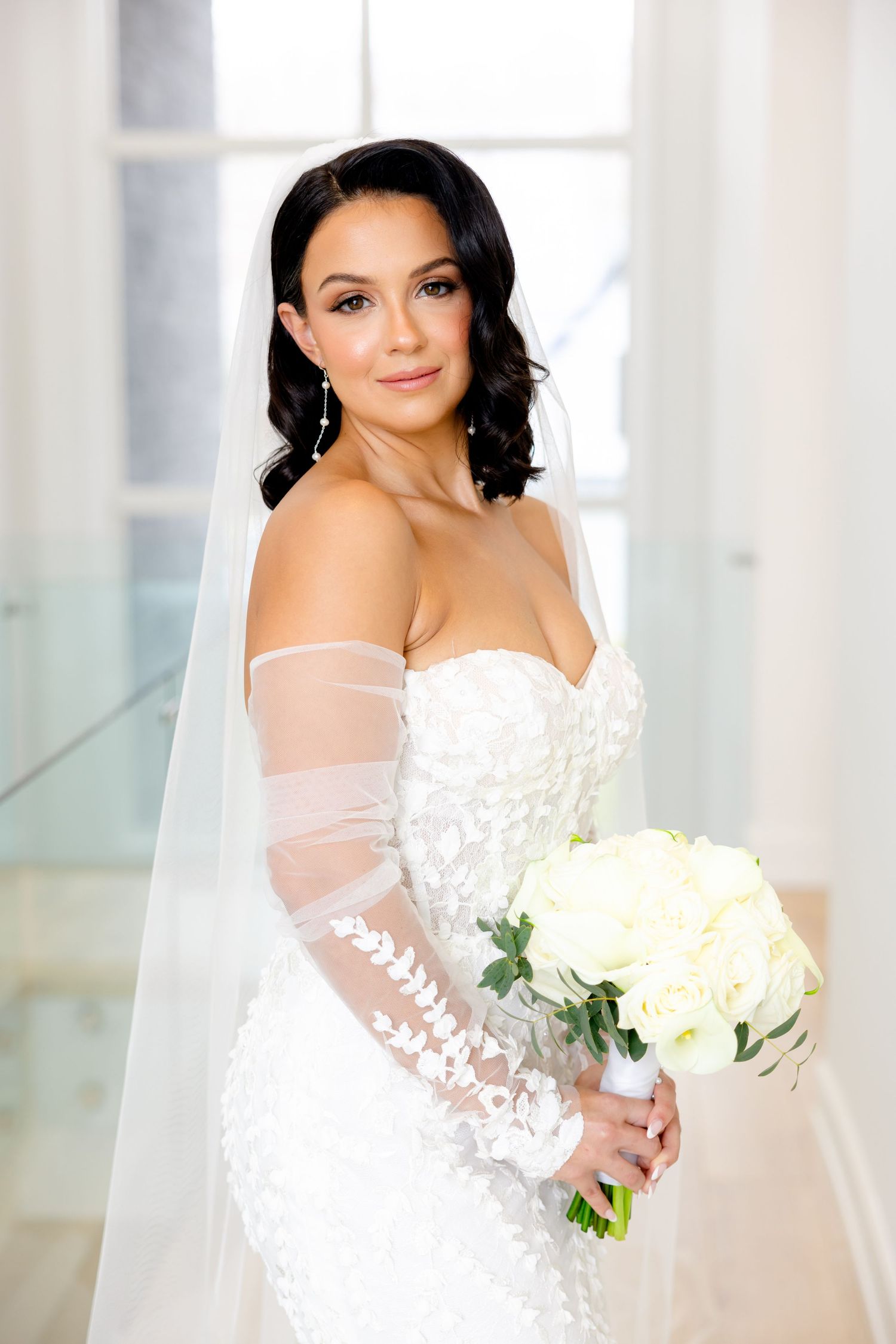 A bridal portrait featuring a strapless lace gown, sheer sleeves, and white rose bouquet.