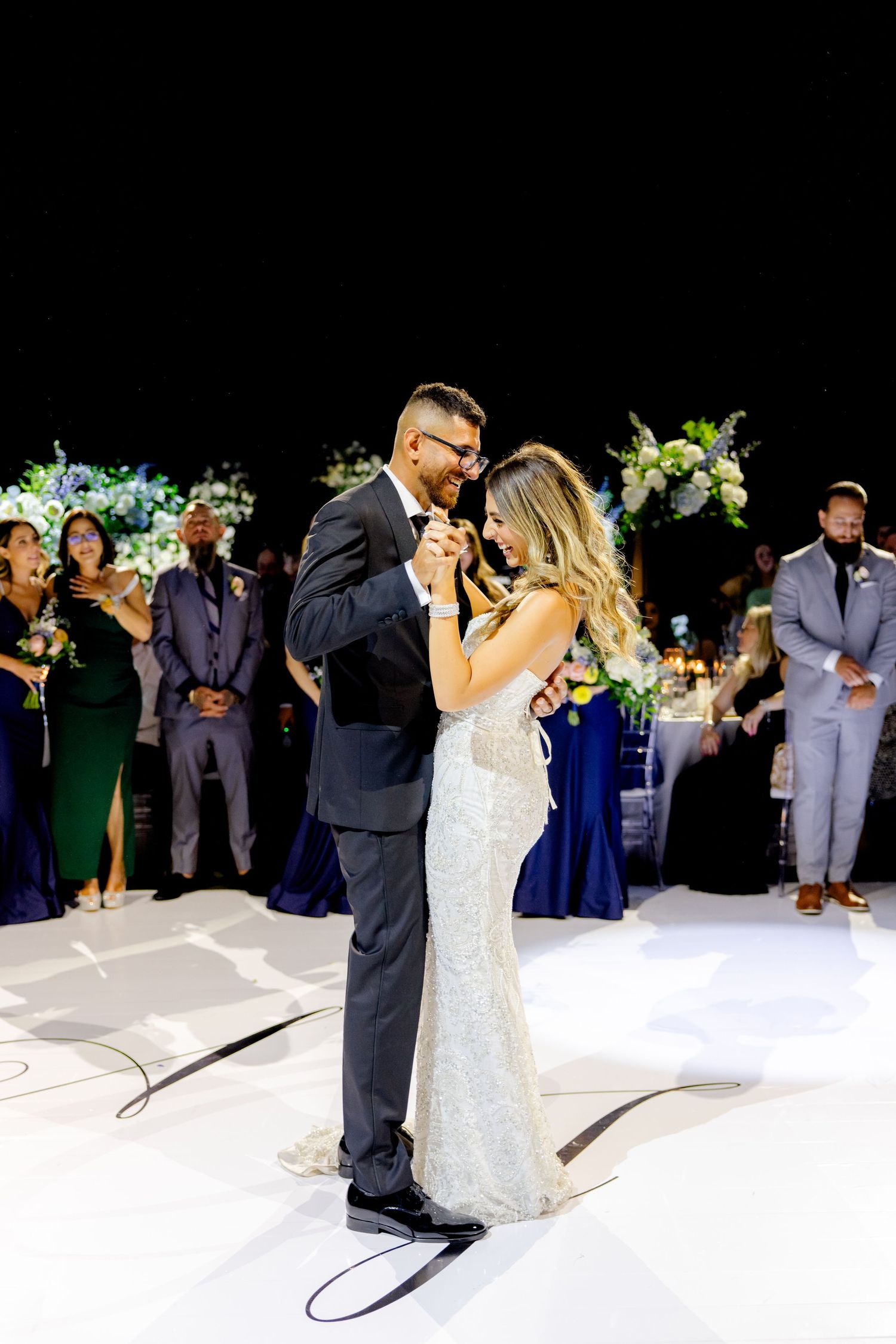 Romantic first dance moment captured with wedding party and floral arrangements in background.