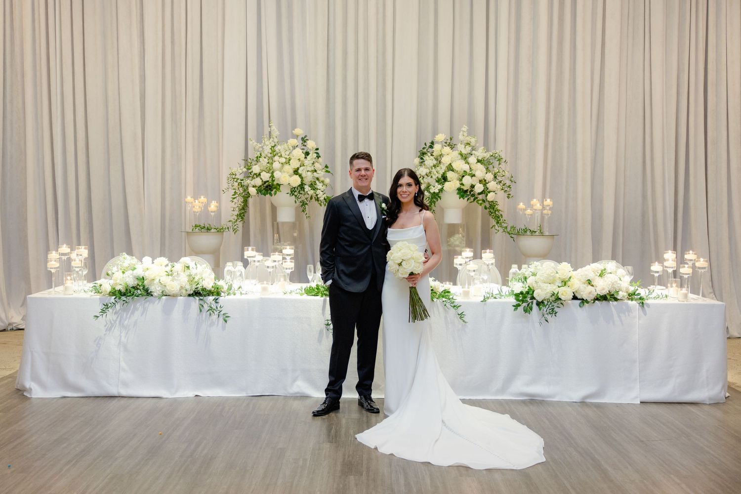 Elegant wedding reception setup with flowing white drapes and lush floral arrangements on white tablescapes.