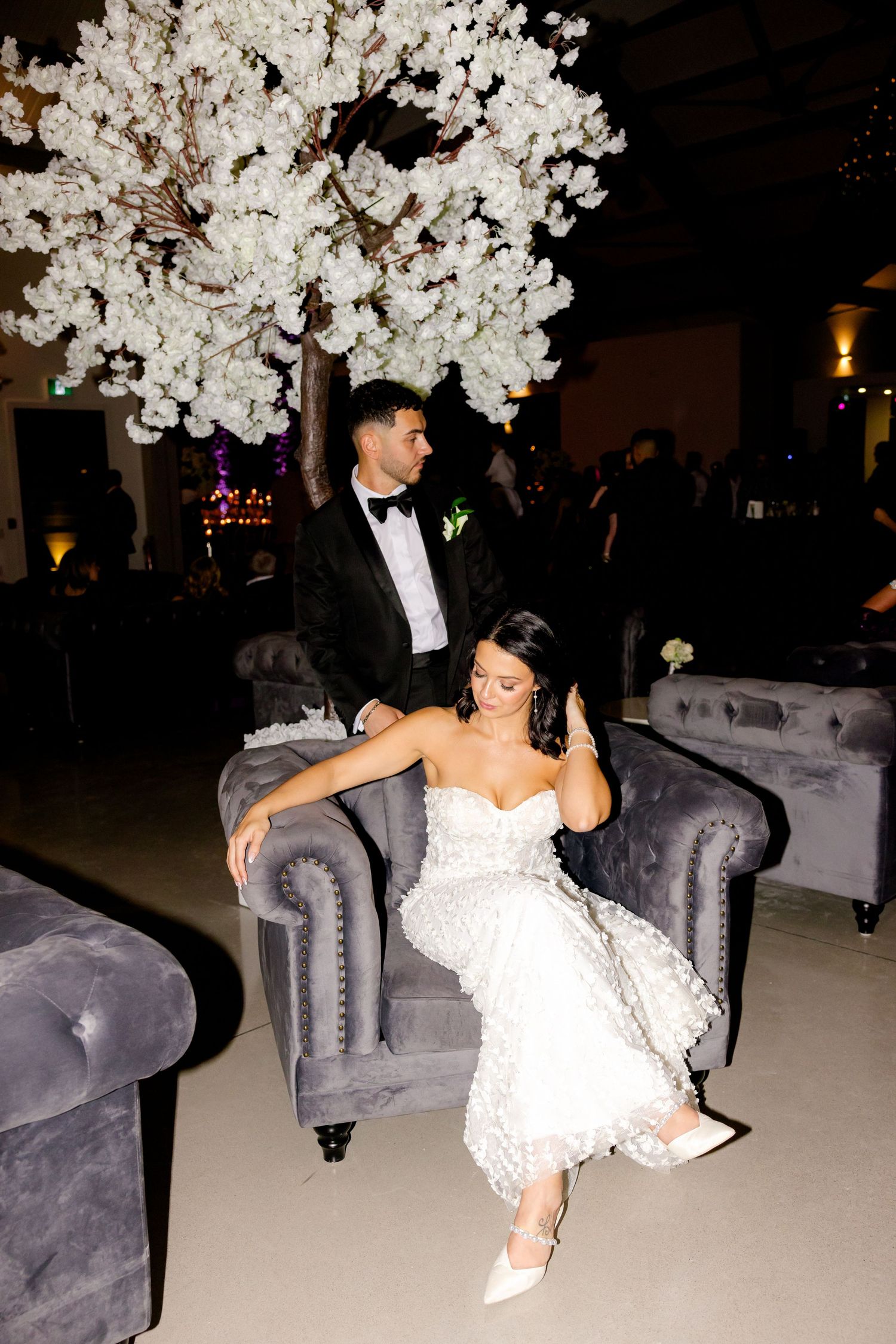 Wedding couple posing on gray velvet couch under white cherry blossom tree decor.