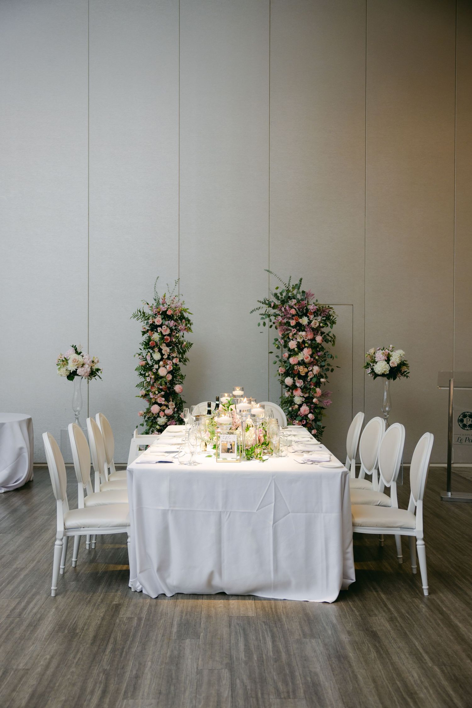 Modern white wedding reception table with floral arch displays and white chairs in minimalist setting.