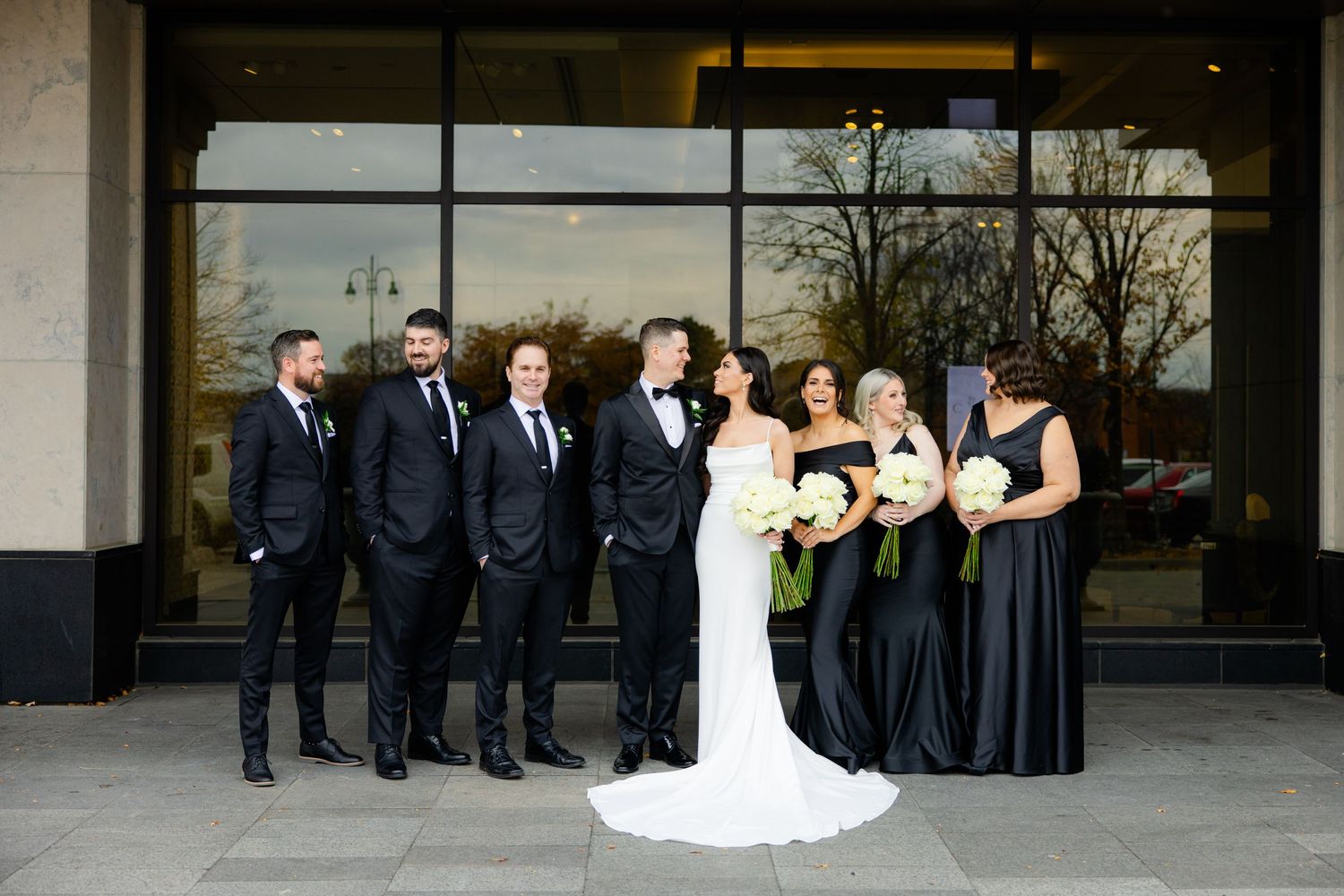 Wedding party in black attire poses together outside modern building with glass windows.